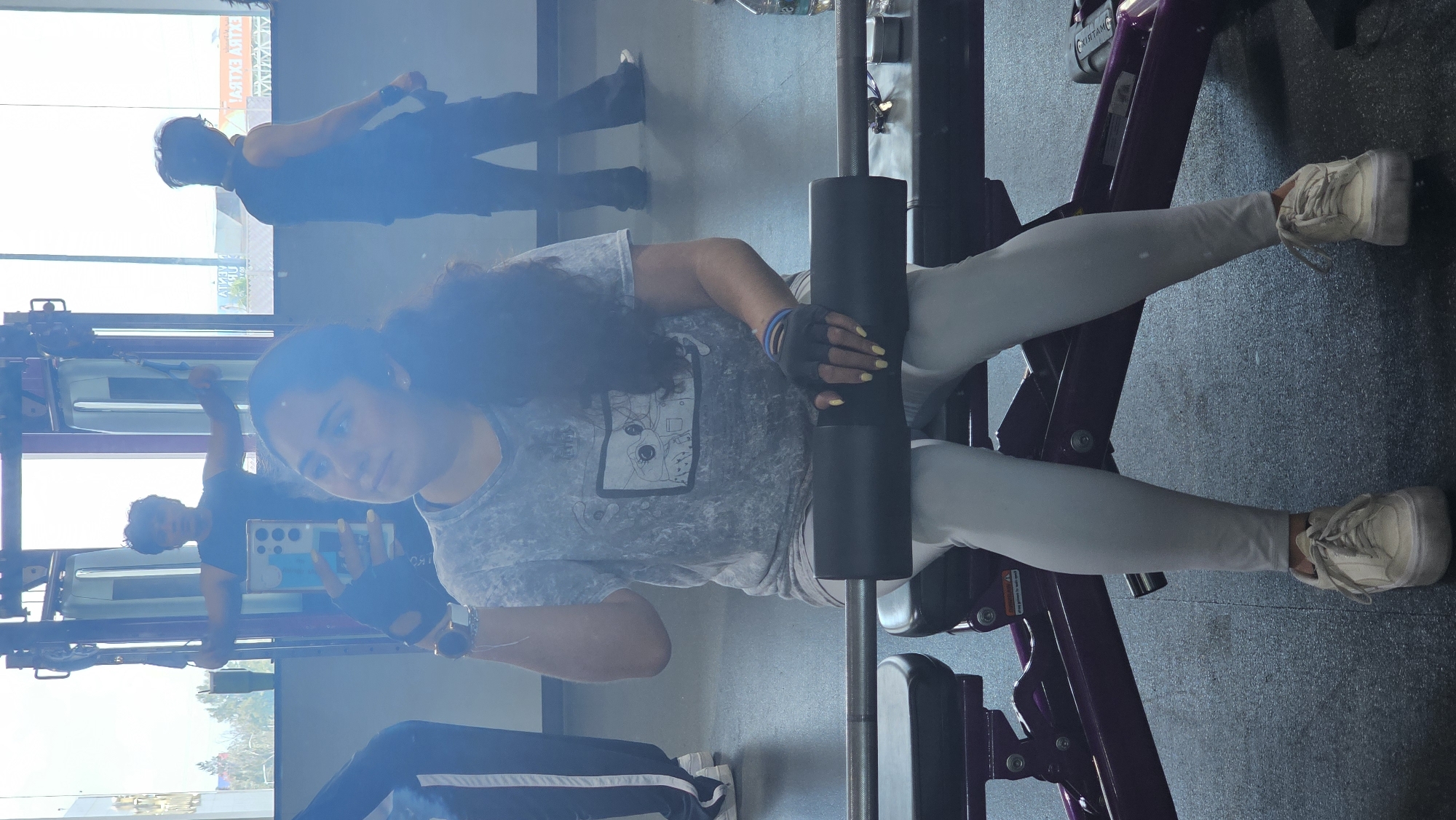 A young woman is captured in a selfie within a gym setting, holding her smartphone (possibly a Samsung S22 Ultra) in her left hand, which reflects part of her face. She is seated on an inner/outer thigh machine, with a black padded bar positioned between her thighs. Her gaze is directed towards the phone's camera. She has long, dark, curly hair and wears a light grey, tie-dye style t-shirt featuring a monochrome graphic print of an anime-like face with indistinct text below, light grey leggings, and light-colored sneakers. She is also wearing black fingerless workout gloves, and her fingernails are painted yellow.
The gym environment features a dark, textured floor and large windows in the background that let in bright daylight. Through these windows, reflections show two other men. One man, wearing a dark tank top and dark pants, is visible standing further back, and another man in a black t-shirt appears to be operating a cable machine. Reflected text on the windows includes "MATRIX" and partially visible, inverted letters that resemble "PLANT FIT" or "PLANET FITNESS." The gym equipment she is using, and other pieces in the background, have purple frames. The scene depicts a daytime workout session in a gym, located in Puebla, Mexico.