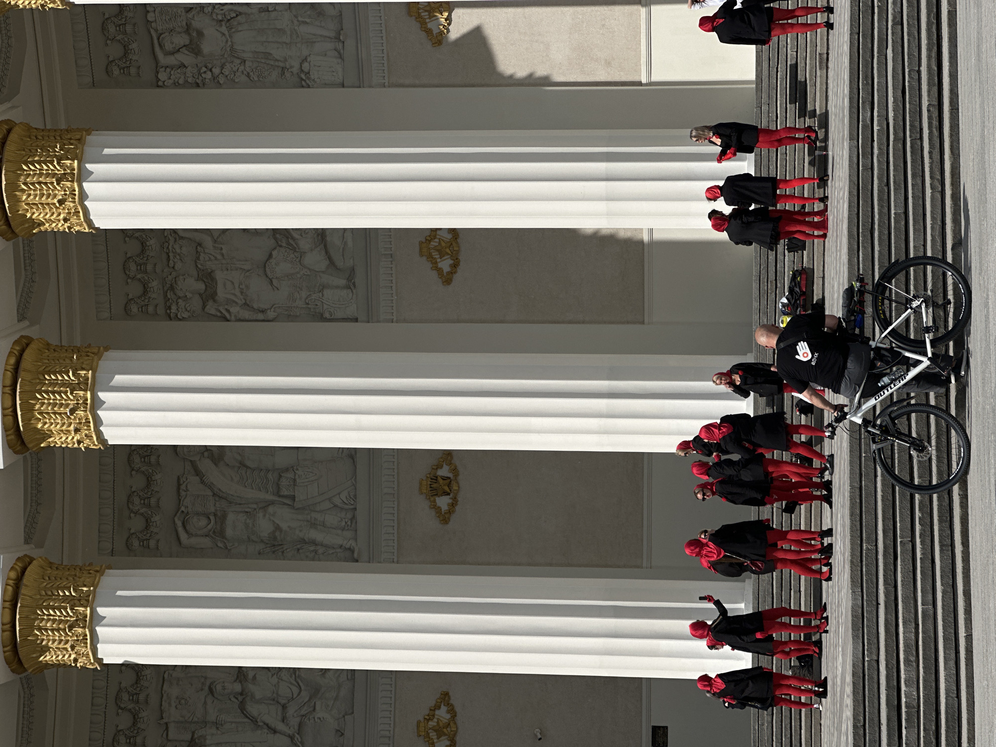 The image shows a building exterior in an unknown location. Three large, white columns with ornate golden capitals stand in the foreground. Relief sculptures are visible on the building's facade between the columns. A group of individuals wearing red leggings, black shoes, and black jackets, with red headscarves, stands on the steps alongside the columns. A man is standing with a white bicycle on the steps.