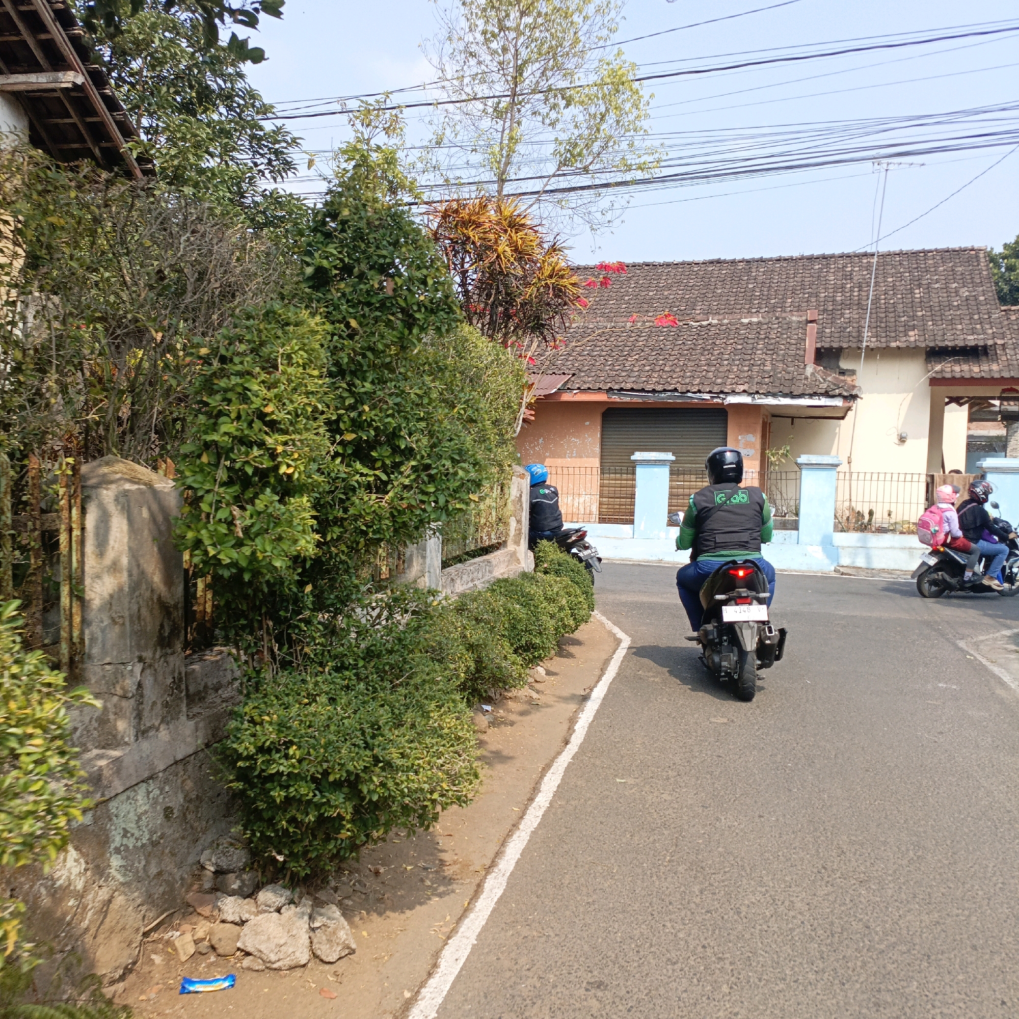 The image depicts a street scene in Salatiga, Indonesia, characterized by a residential area with modest buildings and lush greenery. In the foreground, a gray asphalt road is bordered by a dirt path and a low, manicured hedge. A concrete wall with weathered metal fencing runs alongside the path, overgrown with dense green bushes.
On the road, two motorcycles are visible. The one closer to the viewer is ridden by an individual wearing a black vest with a green "Gojek" logo and a blue helmet, facing away from the camera. The motorcycle itself is black with a visible license plate. Further down the road, another motorcycle is carrying two passengers: a person in the front wearing a black helmet and a jacket, and a child in the back with a pink backpack.
The background features buildings with tiled roofs, some painted in neutral tones and others with brighter accents. The sky is clear blue. Power lines are strung across the upper portion of the image, indicating urban infrastructure. The overall impression is of a typical day in a residential neighborhood in Salatiga.