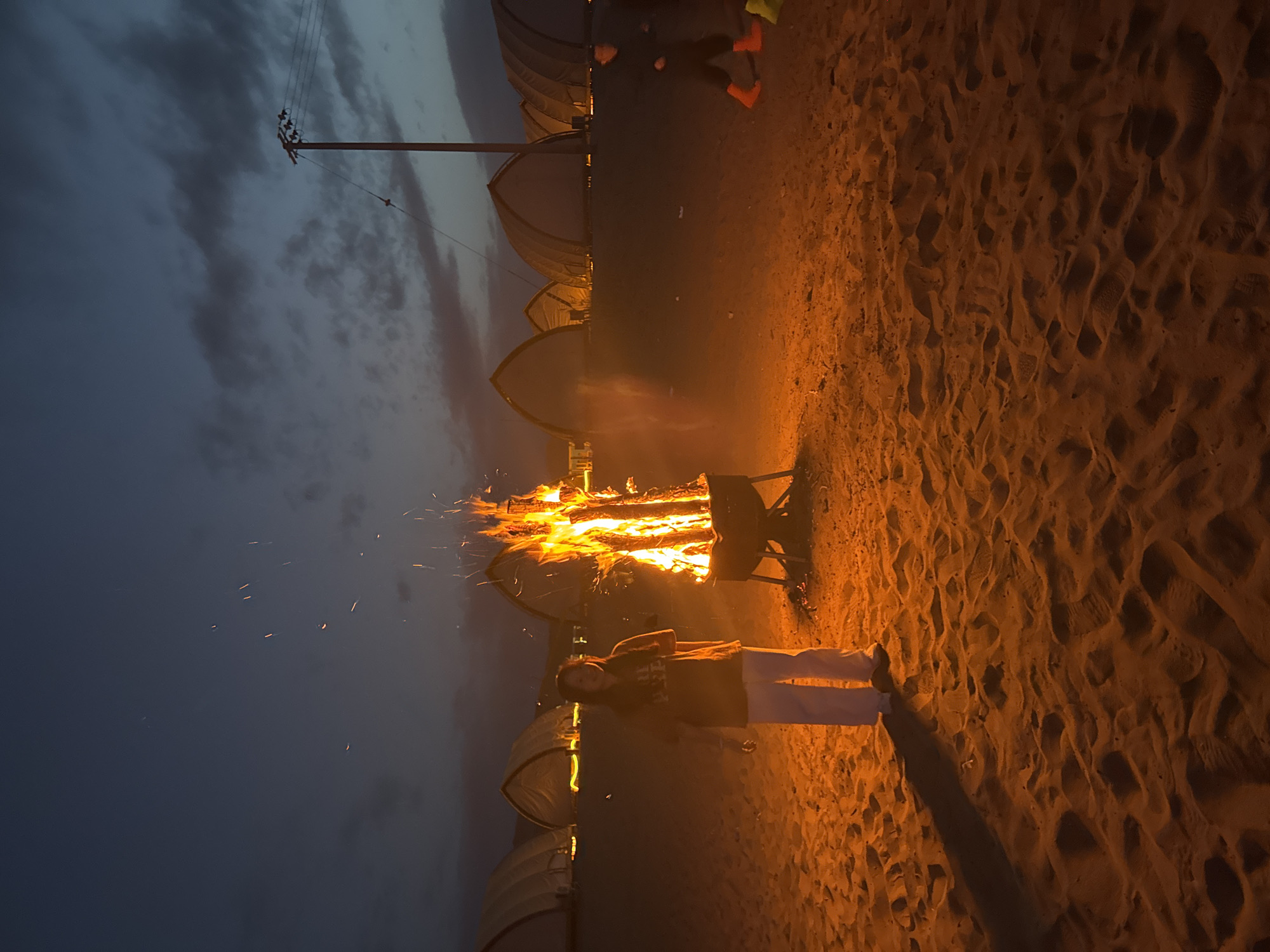 A nighttime outdoor scene features a large bonfire as the focal point, casting a warm glow on the surrounding sand. A person stands near the fire, with their shadow stretching onto the sand. Several dome-shaped structures line the background, illuminated by a dim light. In the distance, a utility pole is visible against a dark, cloudy sky. The scene evokes an outdoor gathering or camping event. The location is Dunhuang City, China.