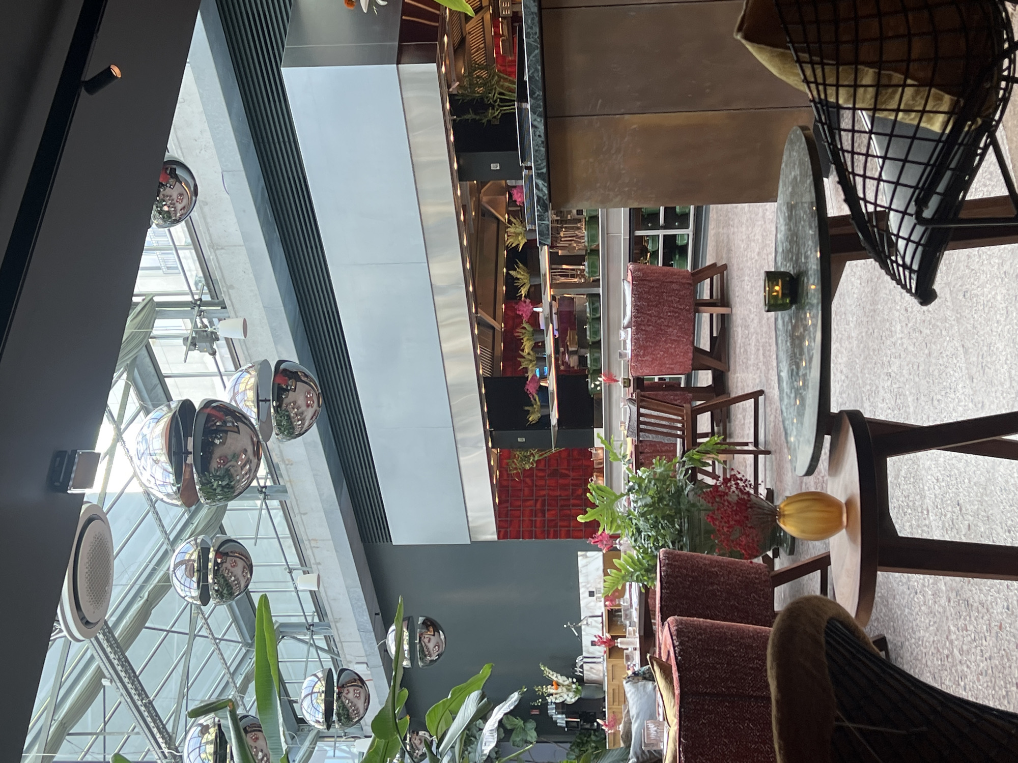 The image shows a well-lit, contemporary restaurant with a high, glass-paneled ceiling, suggesting an indoor-outdoor space, possibly a conservatory or atrium. The interior features modern furnishings including plush red chairs and wooden tables. In the foreground, a small round table holds a delicate amber vase with a sprig of red berries and a small green candle holder with a lit candle. A black wireframe chair with a mustard-colored cushion is partially visible to the right.
Behind the seating area, a long, stainless steel kitchen counter is visible, with stainless steel ventilation hoods above. The kitchen area is characterized by bright lighting and a red tiled backsplash. Various plants and floral arrangements are strategically placed throughout the restaurant, adding a touch of greenery and color to the sophisticated ambiance. Suspended from the ceiling are several large, spherical, mirrored decorative orbs that reflect the surroundings, creating a sense of depth and dynamism. The flooring appears to be a speckled terrazzo. The overall atmosphere is one of upscale dining, with a clean, modern aesthetic. The establishment is the restaurant "Katz Orange" located at Bergstraße 22, 10115 Berlin, Germany.