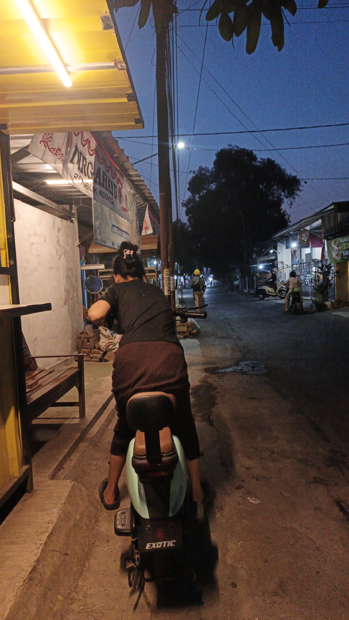 An evening scene on a street in Krimun, Indonesia, features a person seated on a light blue electric scooter with a black seat and backrest. The scooter has the brand name "EXOTIC" visible on its rear. The individual, seen from behind, wears a dark t-shirt and a long, dark brown skirt, with flip-flops on their feet. Their hair is tied up in a bun. They are positioned on the scooter on the side of a paved street, with a concrete sidewalk to their left. To the left of the scooter, a wooden bench is partially visible. Overhead, a yellow awning with fluorescent lighting extends from a building. Banners with Indonesian text, including "DIRGAHAYU" and "REPUBLIK INDONESIA," are displayed on the building facade. The sky is a deep blue, indicating twilight. In the background, other people and scooters are present on the street, and buildings line the right side of the road. A prominent utility pole with numerous wires stands near the center of the frame. A dark tree silhouette is visible against the sky.
