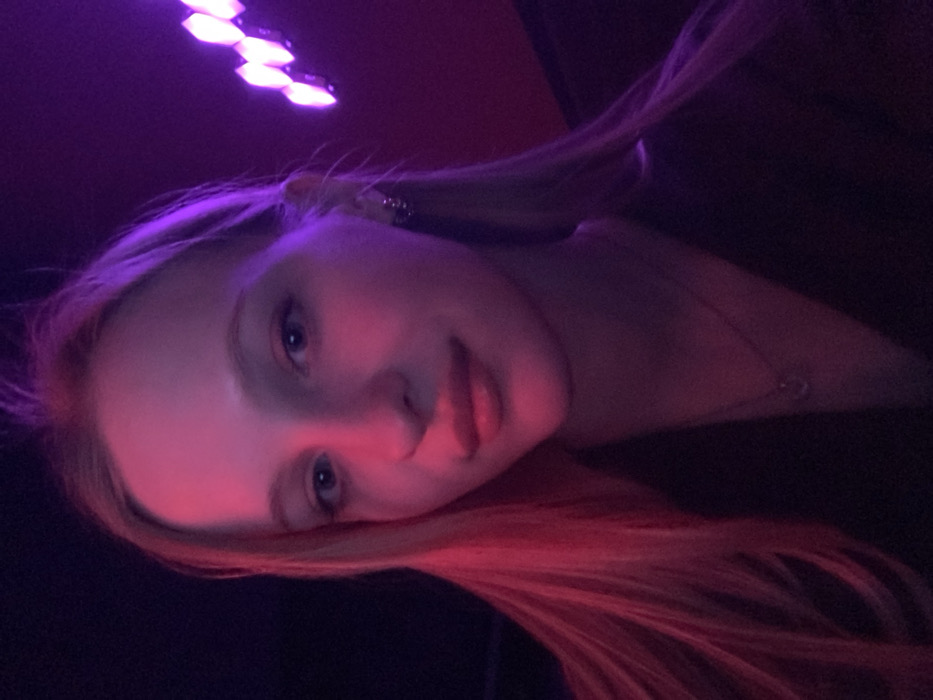 A young woman with long blonde hair and light-colored eyes is depicted in a medium close-up, facing forward with a neutral expression. Her features are illuminated by a dual-color artificial light source: the left side of her face and hair is bathed in a strong pink-red light, while the right side is lit by a vibrant purple hue. She wears a dark top, a thin necklace with a small, light-colored, oval pendant, and small silver-toned hoop earrings on her right ear. The background is dark and out of focus, revealing a cluster of illuminated, geometric purple light fixtures in the upper right quadrant. This setting suggests an indoor entertainment venue, such as a nightclub or lounge. There are no identifiable visual cues to determine a precise location within Kyiv, Ukraine.