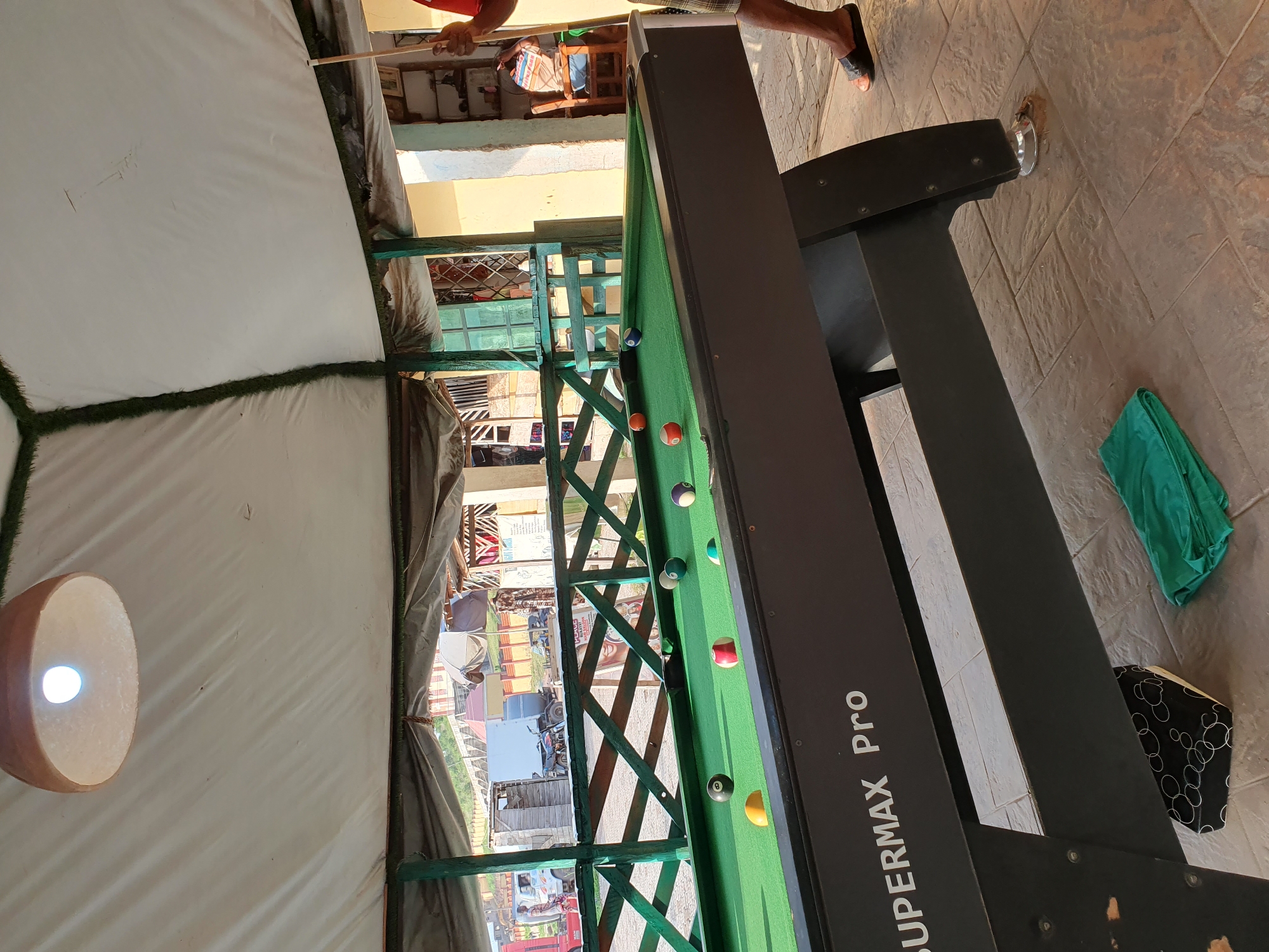 An outdoor pool hall in Lokoja, Nigeria, is captured in a medium shot. The scene is set under a white canopy with a decorative grass trim. A pool table, marked "SUPERMAX Pro," dominates the foreground, its green felt surface populated with several billiard balls. A person, partially visible, stands to the right, holding a pool cue, suggesting an ongoing game. The background reveals a street scene with vehicles and buildings, indicating a lively urban environment. The floor is paved with textured tiles, and a green cloth lies on the ground near the pool table.