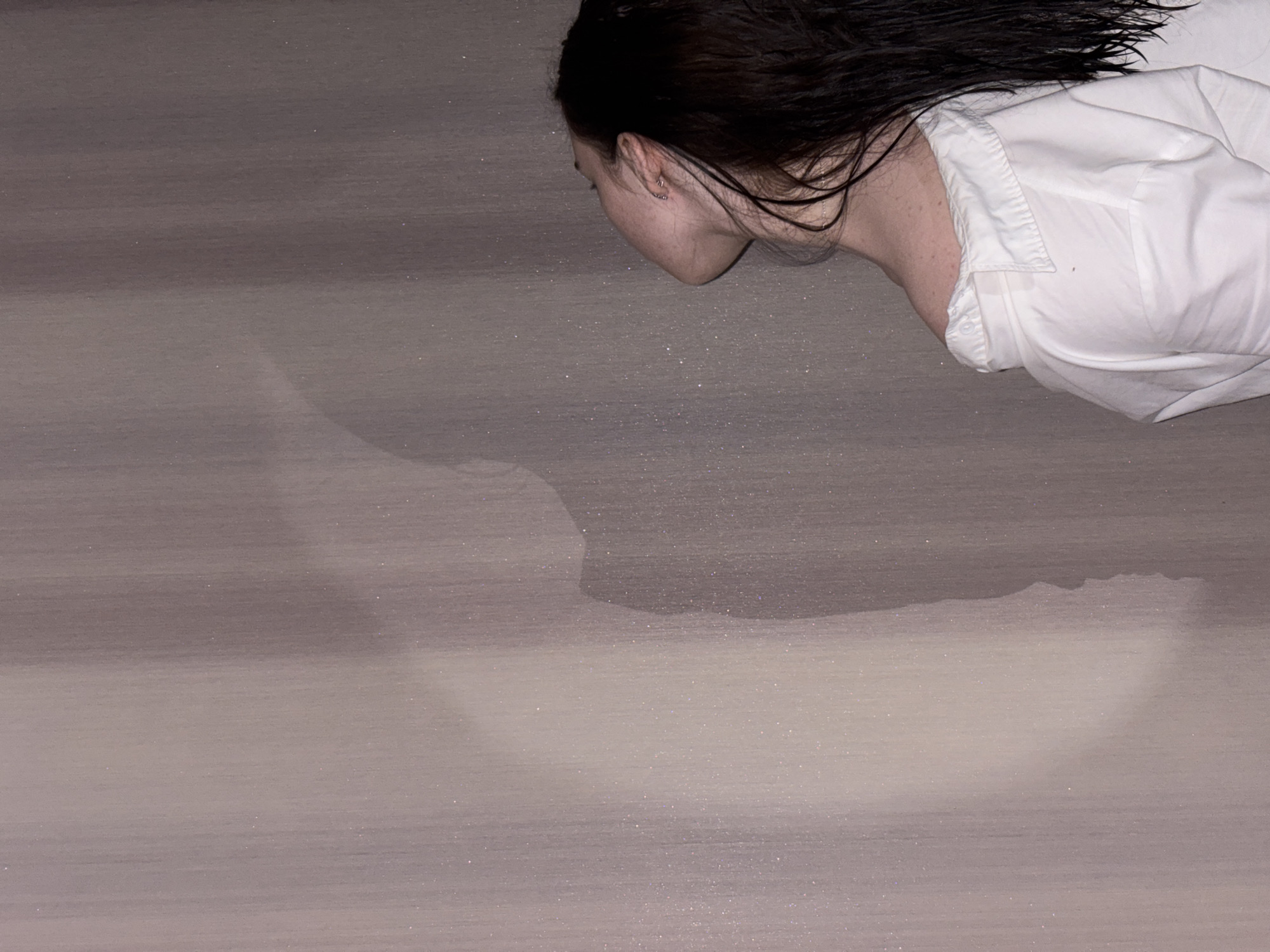 A stylized, horizontal portrait depicts a person, likely female, with dark, wet hair, positioned against a subtly textured, monochromatic background. The individual is seen in profile from the upper right portion of the frame, with her head tilted downwards, obscuring her face from direct view. She wears a white collared shirt, and small, metallic earrings are visible on her ear. Her dark hair appears damp, splayed around her head.
The background is a gradient of muted grey and brown tones, displaying soft horizontal striations that suggest a painted or textured surface. Numerous tiny, sparkling particles, resembling glitter or illuminated dust, are scattered across this surface, creating a subtle shimmering effect. These particles are noticeable throughout the background and around the individual's head and neck.
The lighting is soft and diffused, creating an even illumination that accentuates the texture of the background and the details of the subject's hair and clothing. The person's posture and downward gaze lend the image a contemplative or introspective mood. There is no explicit activity or event occurring, indicating a posed or artistic composition. The image provides no specific time of day, weather conditions, or visual location cues within the frame itself. No text is visible. The location context for this media is Perm, Russia.