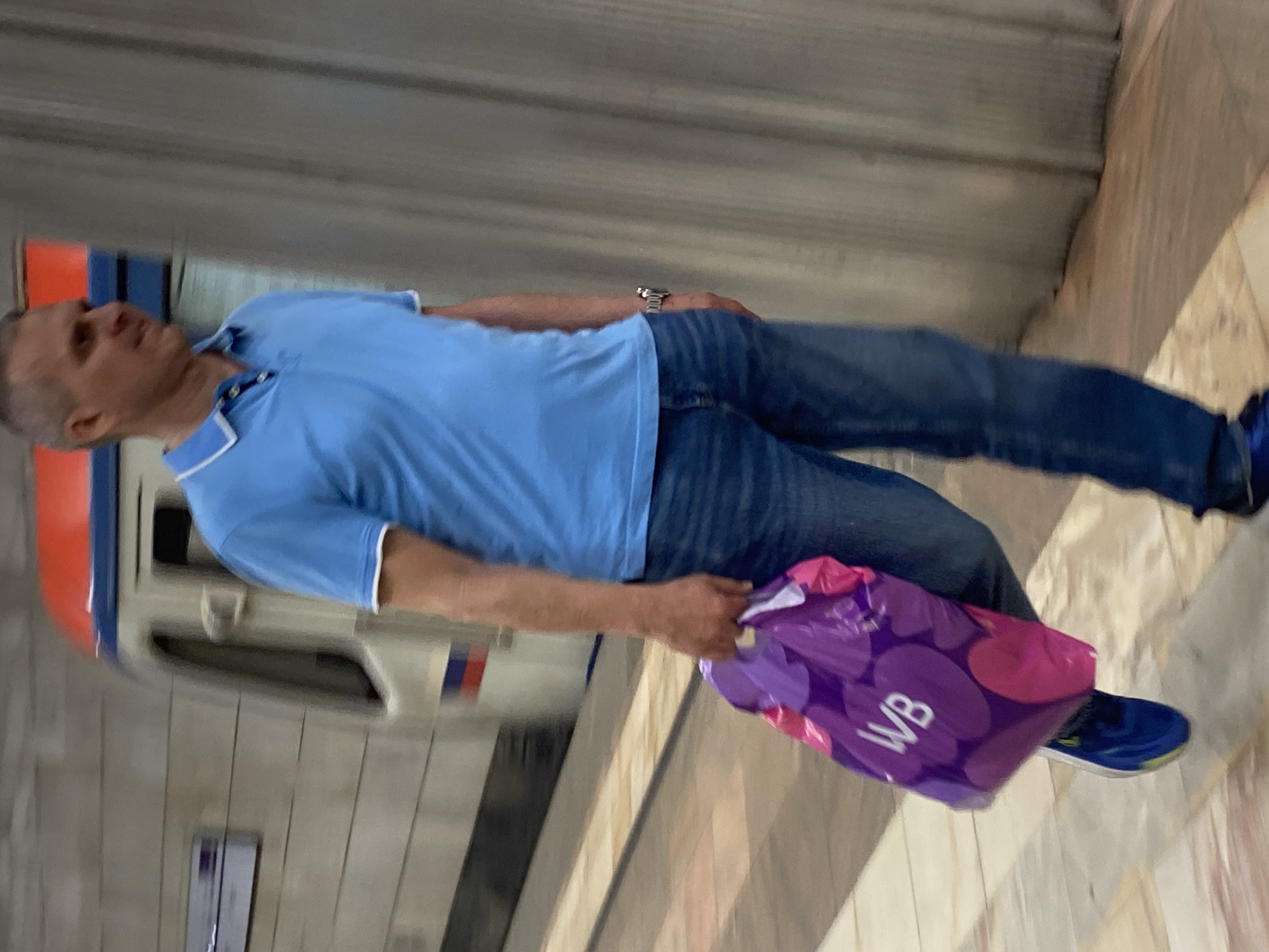 A man is walking on a platform in a metro station in Khimki, Russia. He is wearing a blue polo shirt, blue jeans, and blue and yellow sneakers. He is carrying a purple shopping bag with "WB" written on it in white letters. A train is stopped at the station behind him, its windows blurred with motion. The station has concrete walls and a tiled floor.