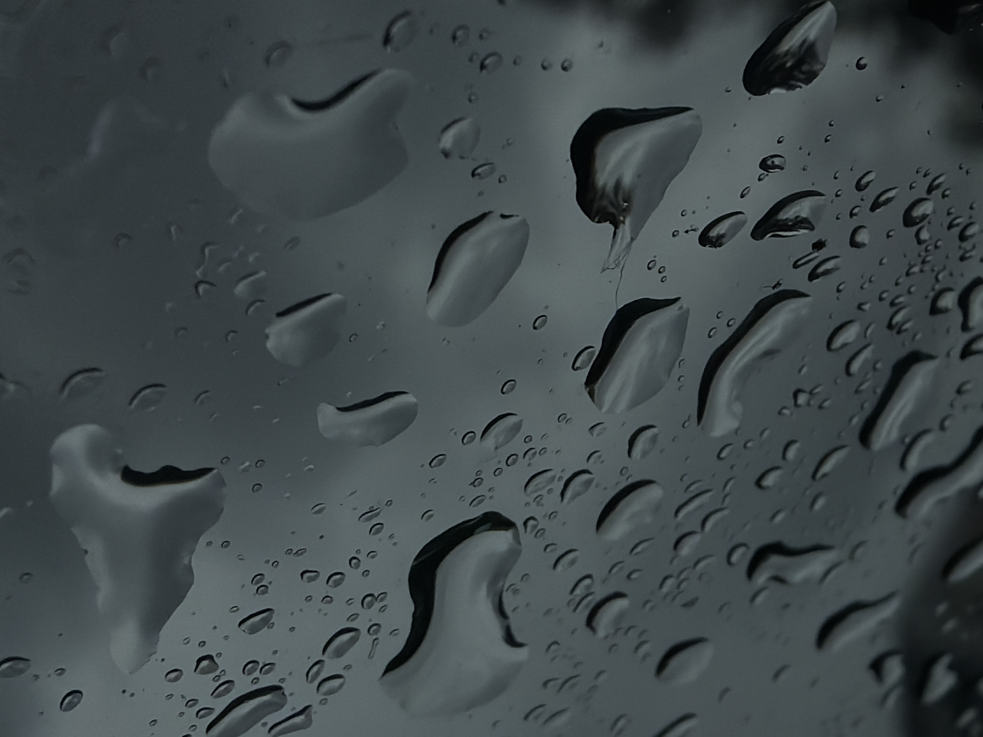The image depicts raindrops on a window, with an overcast sky visible through them. The raindrops vary in size, from small droplets to larger, irregularly shaped formations. The lighting is dim and the color palette is monochromatic, predominantly shades of gray. No people, subjects, or objects can be identified beyond the raindrops and the diffused background sky. The location in Naberezhnye Chelny, Russia, cannot be verified from this image.