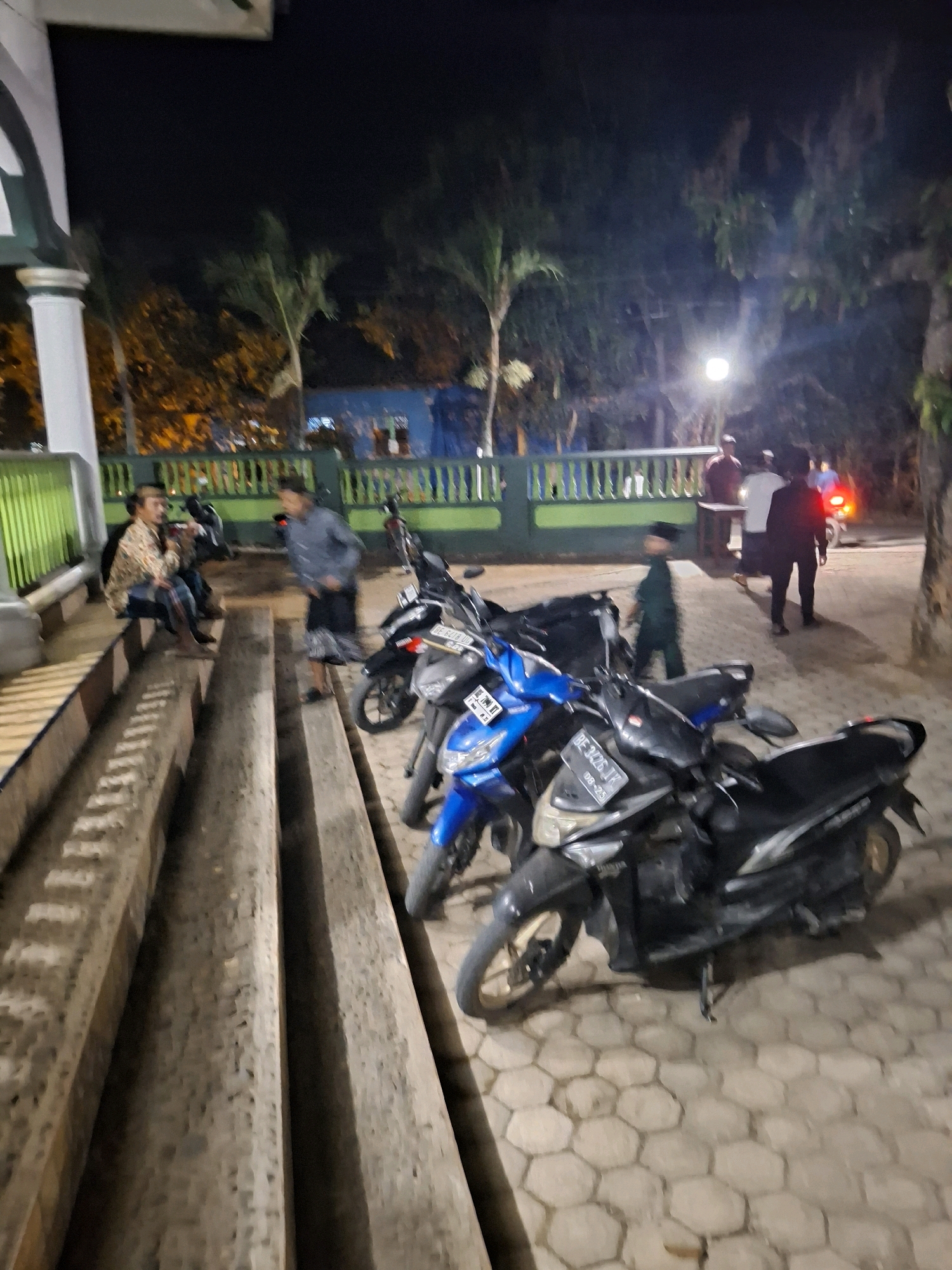 The image depicts an outdoor scene at night, possibly in Yogyakarta, Indonesia, near a building with white pillars and green fencing. Several people are present, and a row of parked motorcycles is a prominent feature. In the foreground, three scooters are parked on hexagonal paving stones. The closest scooter is black, followed by a blue one, and then another black scooter. Further back, more motorcycles are visible.
On the left side of the image, two men are seated on concrete steps. One man, wearing a patterned shirt and jeans, is facing the other man, who is wearing a dark shirt and a sarong. The man in the sarong appears to be speaking. Nearby, another person is walking away from the seated individuals, wearing a blue shirt.
In the background, beyond the green fence, a group of people are standing near a lamppost. One man in a dark shirt and trousers stands out. There is also a red taillight visible, suggesting a vehicle or motorcycle in motion further in the background. Trees and foliage are visible, illuminated by artificial light, creating a nocturnal atmosphere. The overall scene suggests a social gathering or a resting spot outside a building.