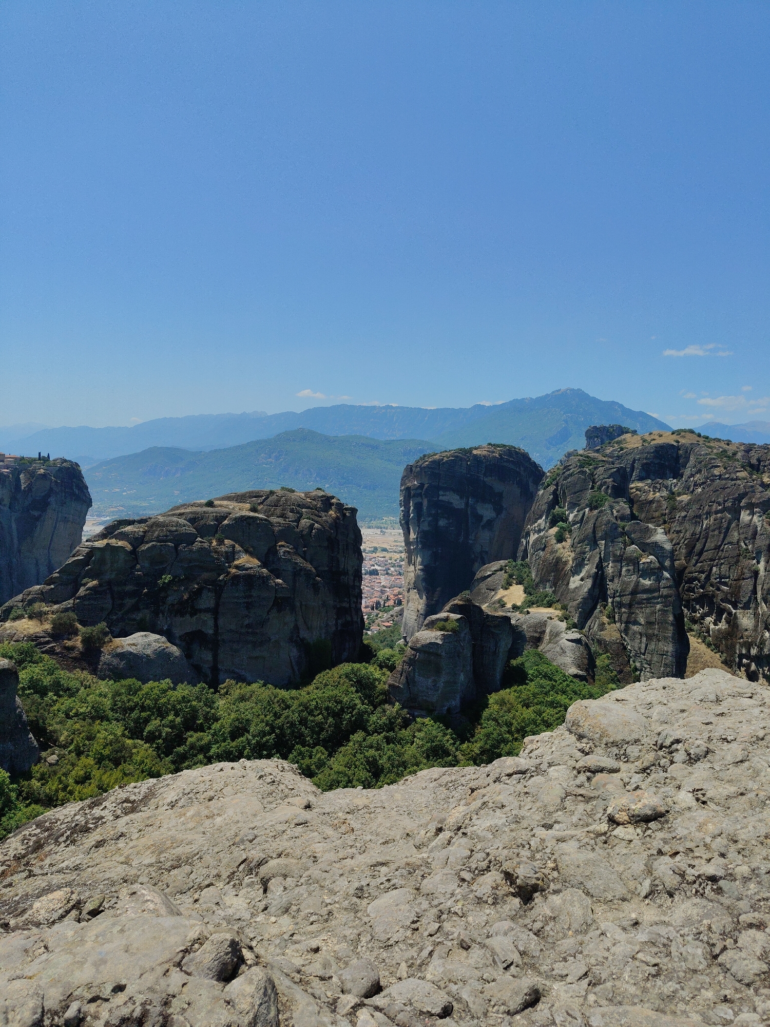 This is a vertical, medium-shot, eye-level photograph of the rock formations of Meteora in Kalambaka, Greece. The sky is a clear, vibrant blue, taking up the top third of the frame. Below the sky are several towering, imposing rock pillars with sheer cliff faces. These formations are dark gray and textured, with some vegetation clinging to their surfaces. The foreground is dominated by a rough, gray rocky surface, providing a textured foreground. In the middle ground, between the rock formations, a town is visible with red-tiled roofs and some white buildings. Lush green trees and foliage grow at the base of the rock formations and in the valley below. In the distance, rolling hills and mountains fade into a hazy blue landscape. The scene depicts a natural landscape with human settlements integrated into it. There are no people or animals visible in the frame. The lighting suggests it is daytime, with bright sunlight casting shadows on the rocks.