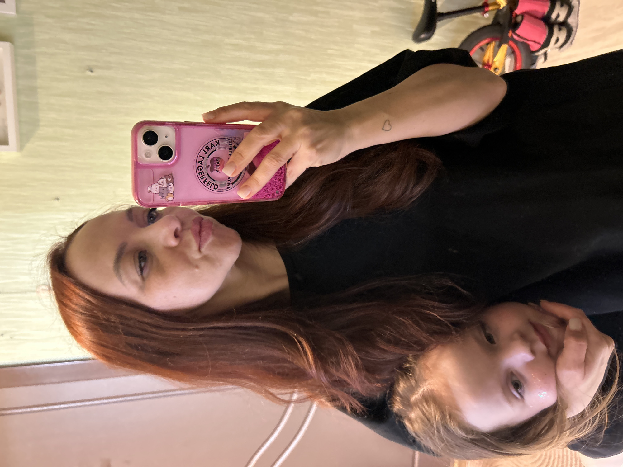A woman with reddish-brown hair and a small heart tattoo on her left wrist holds a pink iPhone case with a Karl Lagerfeld design, taking a selfie with a young child. The child has fair skin and blonde hair. The background includes a wall, part of a child's tricycle, and a door, suggesting an indoor environment, possibly a home. The city of Chaikovsky, Russia, is not discernable from the provided image.
