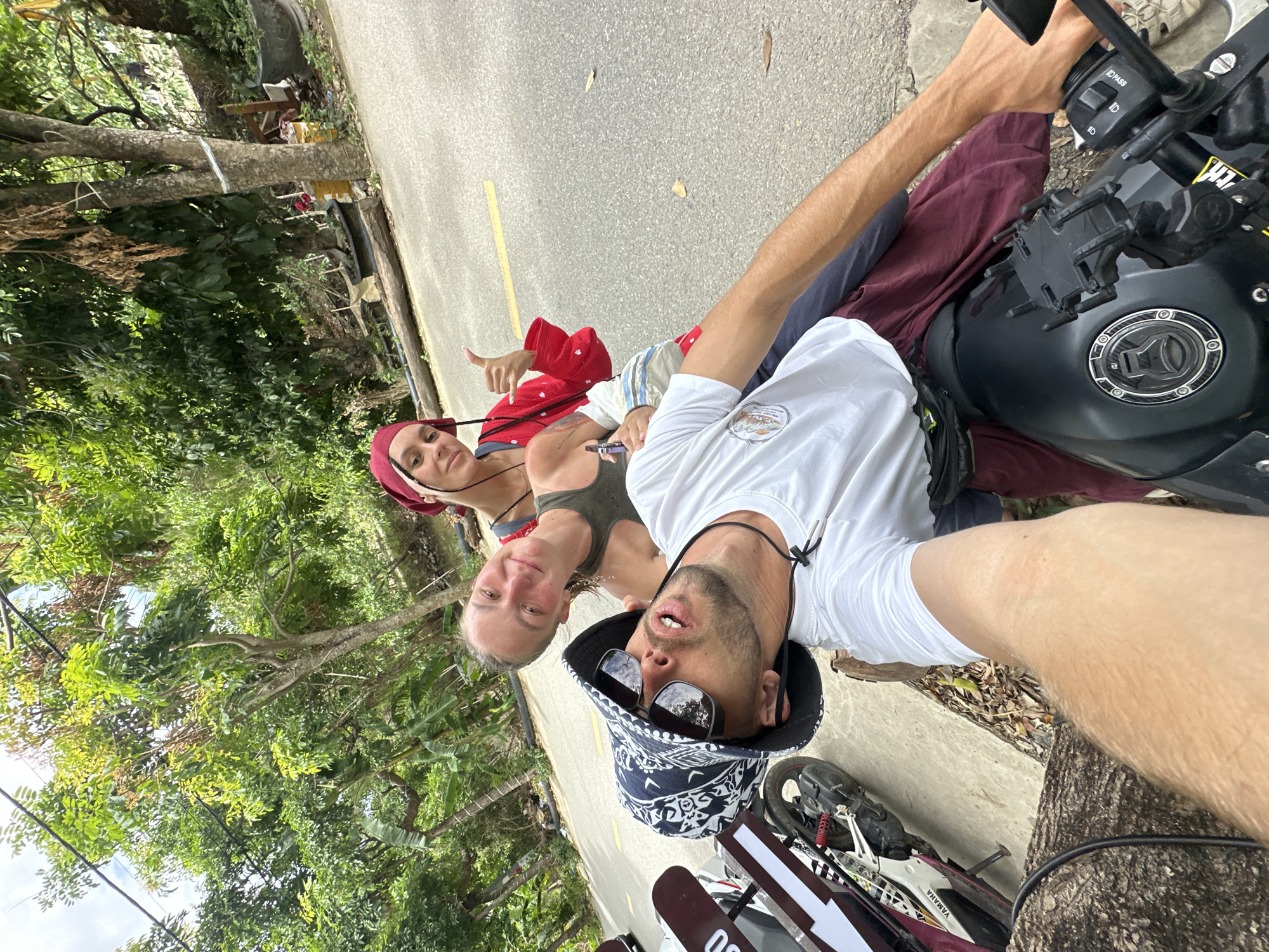 Three individuals are posing for a selfie on a motorcycle on a paved road in Nam Hoa Lư Ward, Vietnam. The person in the foreground, wearing a white t-shirt, a patterned bucket hat, and sunglasses, is leaning back and holding the camera. A second person, wearing a green tank top and a red bandana, is seated behind the first person, smiling and giving a thumbs-up. A third person, wearing a red jacket and a red bandana, is partially visible behind the second person. The background shows lush greenery, trees, and a body of water. A white and red Yamaha motorcycle is parked to the left of the frame.