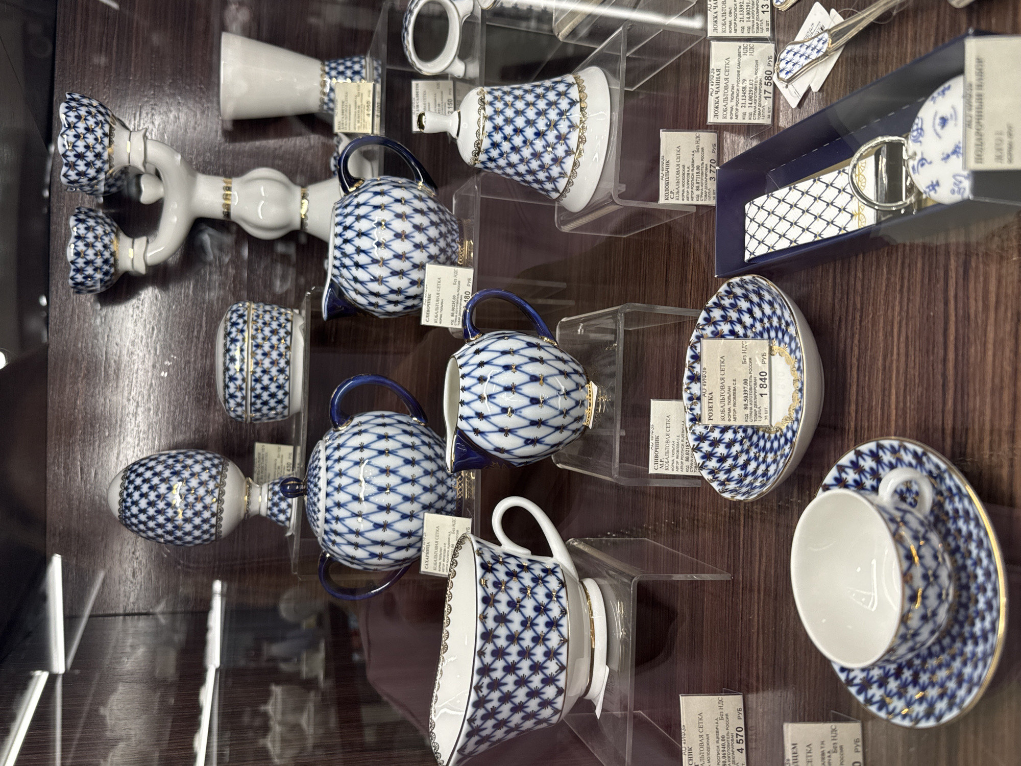 The image shows a collection of porcelain items with a blue and white lattice pattern and gold accents, likely Gzhel porcelain, displayed in a glass case. These items include teapots, cups and saucers, a bell, a candlestick, a sugar bowl, and spoons. The prices are indicated on labels in Russian rubles. The origin of the porcelain is specified as Russia. The scene appears to be inside a store or a museum, where these products are showcased for sale or exhibition. The Russian text on the labels suggests this image was taken in a location in Russia, possibly the city of Saint Petersburg, Russia.