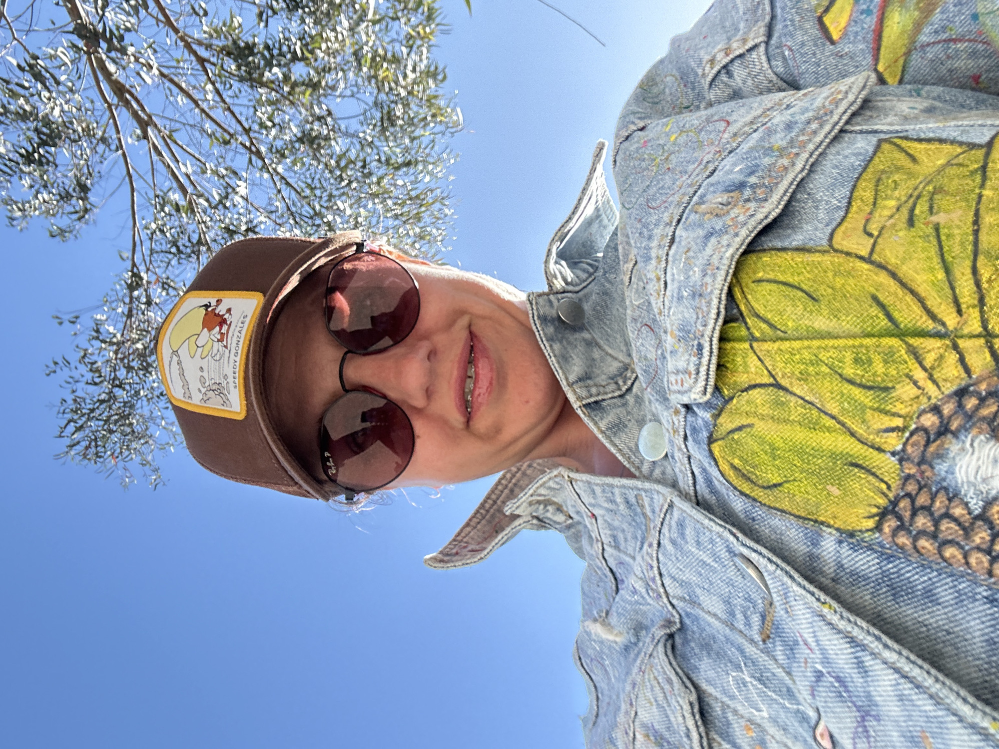 A low-angle shot captures a smiling woman wearing a brown baseball cap with a "Speedy Gonzales" patch, round sunglasses, and a light denim jacket adorned with a painted sunflower design. Her teeth with braces are visible. Branches of a tree with small leaves are visible in the upper left corner, while a clear blue sky fills the background. This scene does not provide information to determine its location in the city of Sochi, Russia.