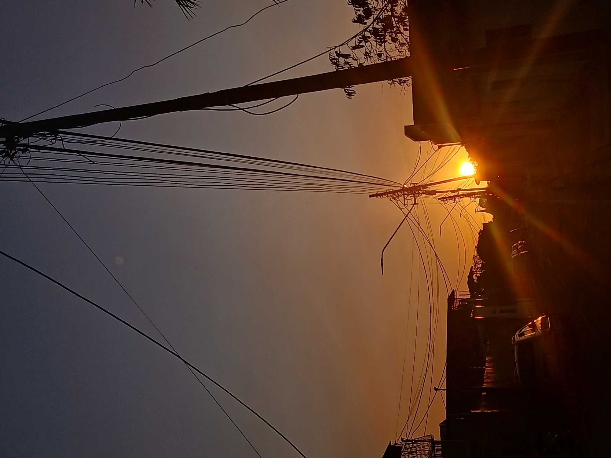 The image shows a street scene in Juan Griego, Venezuela at sunset. The sun is positioned behind buildings on the right side of the frame, creating lens flares. A power pole stands on the left side of the frame, with numerous electrical wires extending across the sky. The sky transitions from dark blue on the left to orange near the sun. A vehicle is partially visible parked along the street on the right side of the frame.