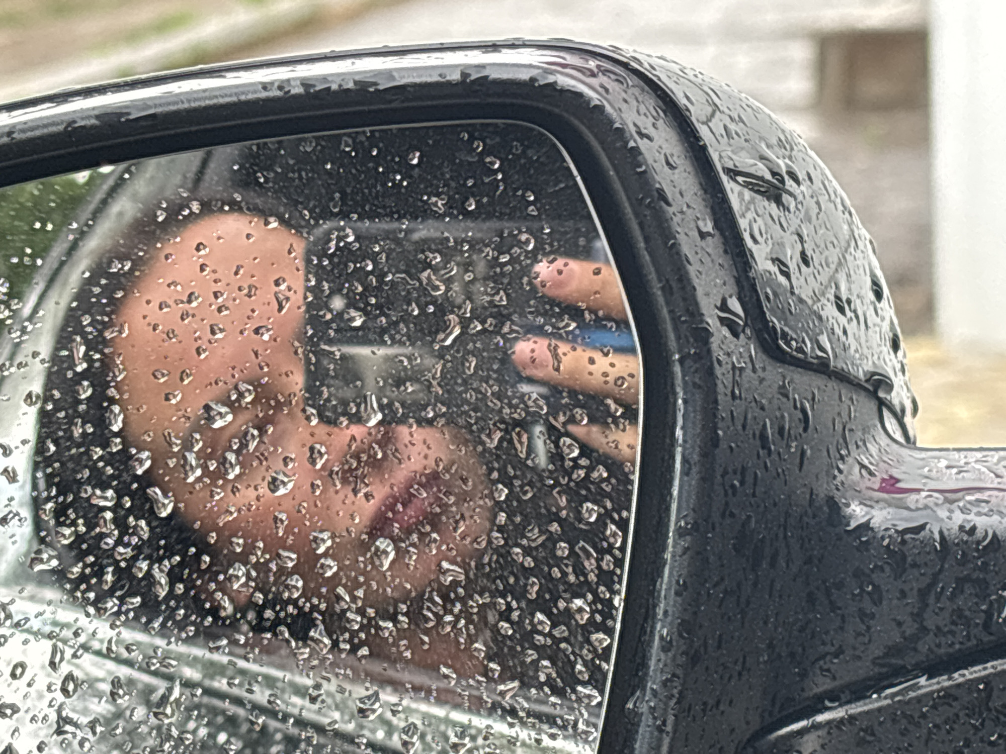 The image depicts a close-up reflection in a car side mirror covered in raindrops. The reflection shows a person holding a smartphone to take a picture. The environment appears to be outdoors during or after a rain shower, with a blurry background suggesting a residential or urban area. The scene's location is not identifiable.