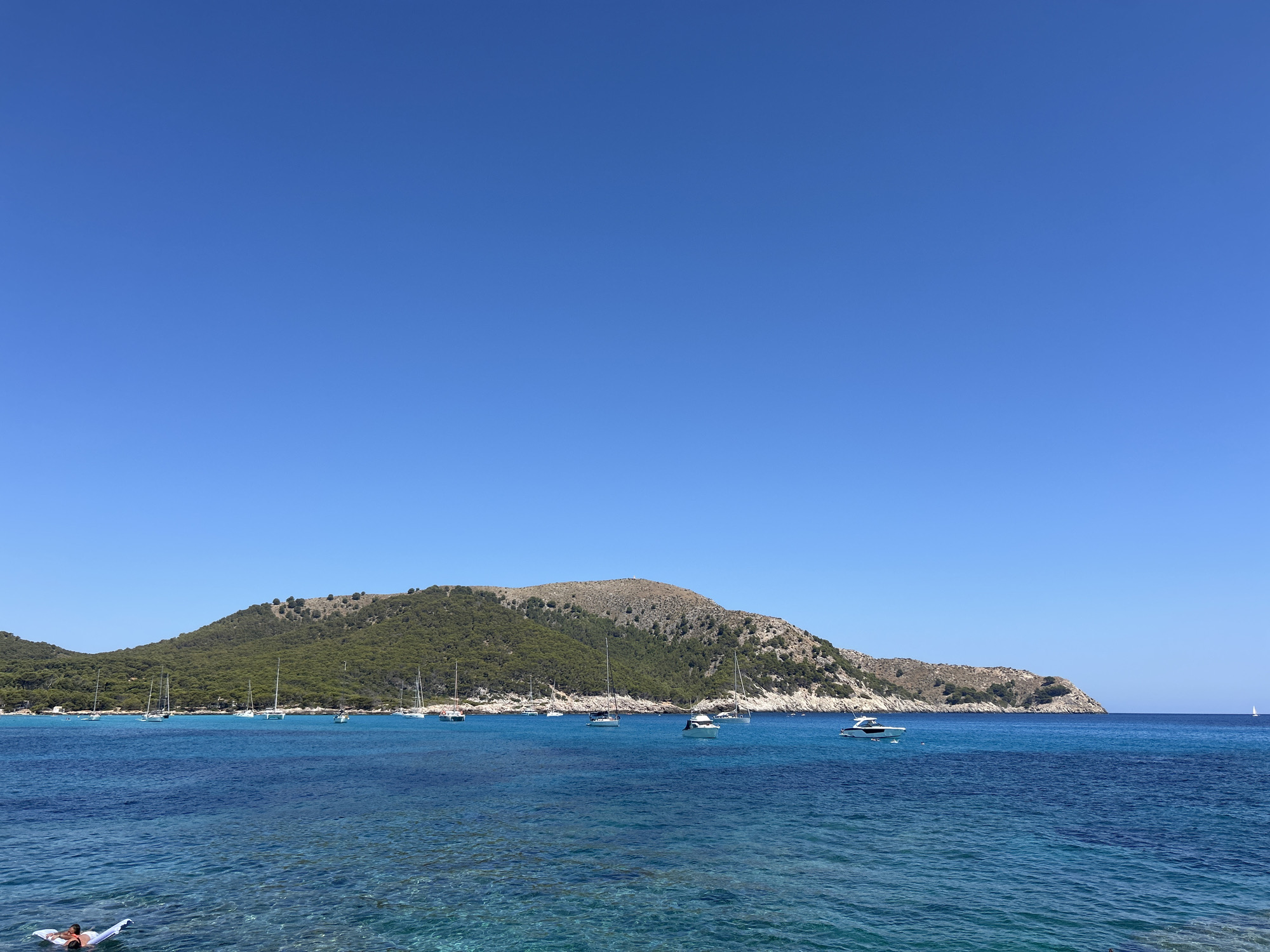 The image shows a panoramic view of a bay in Cala Lliteres, Spain, on a clear, sunny day. The sky is a vibrant, cloudless blue, taking up the top half of the frame. Below the sky, the deep blue sea stretches across the entire width of the image, with varying shades of turquoise and azure near the shore. The water is calm, with gentle ripples reflecting the sunlight.
In the foreground, a person is seen floating on an inflatable raft. They are partially submerged in the water, appearing to be relaxing.
Anchored in the bay are numerous sailboats and a few motorboats, their white sails and hulls stark against the blue water. The boats are scattered across the middle ground of the image, suggesting a popular anchorage.
Behind the bay, a lush, green, and hilly landscape rises up. The hills are covered in dense vegetation, primarily pine trees and shrubs, with some rocky outcrops visible. The contours of the land suggest a Mediterranean environment. The overall impression is one of a serene coastal landscape, ideal for leisure and recreation.
