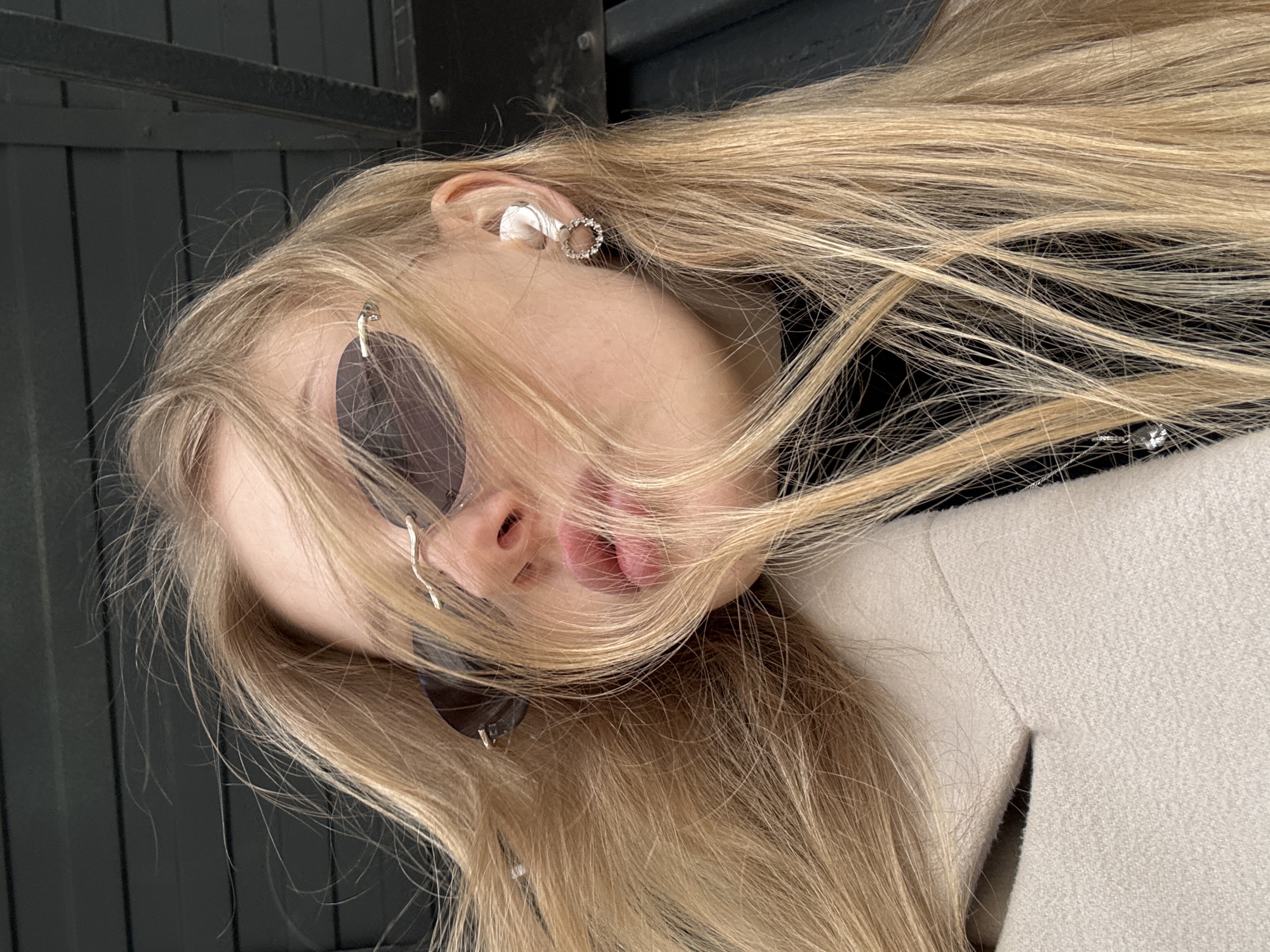 A blonde woman is depicted from a low, tilted perspective. She wears sunglasses and an earring, and has an earpiece in her ear. She is wearing a cream-colored coat. The background is a dark, slatted or paneled surface. The location is presumed to be Perm, Russia.