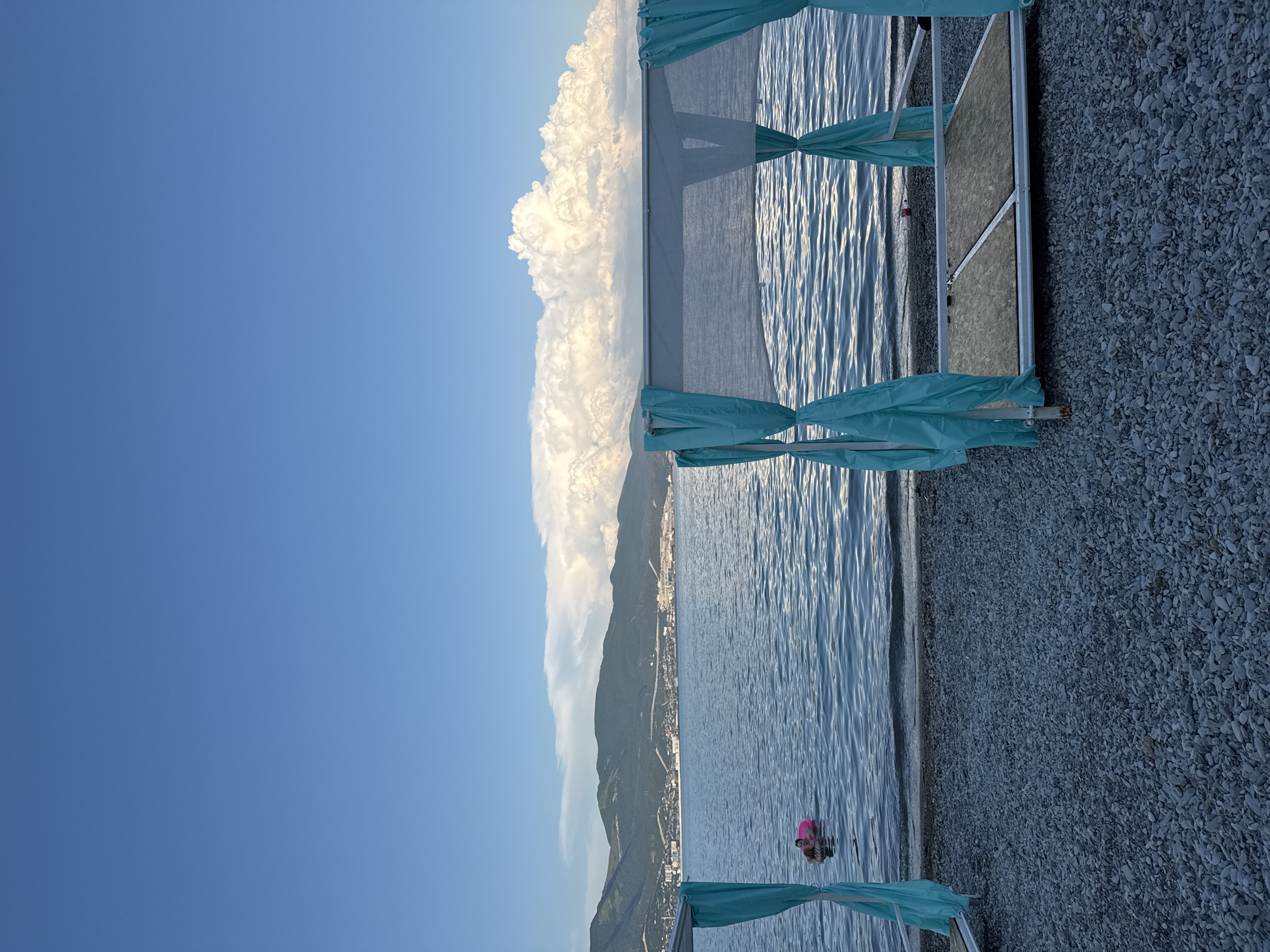 The image depicts a serene beach scene in Neberdzhaevskaya, Russia. In the foreground, a gravel beach is occupied by a white canopy with turquoise curtains, suggesting a private lounging area. The canopy has a wooden platform for seating or reclining. In the middle ground, the calm blue sea gently laps against the shore. A person is visible in the water, wearing a pink inflatable ring and seemingly enjoying a swim. In the background, a picturesque mountain range forms a majestic backdrop to the coastal town, which is dotted with buildings and structures. The sky above is a clear, vibrant blue, with large, fluffy white clouds adding depth and texture to the horizon.