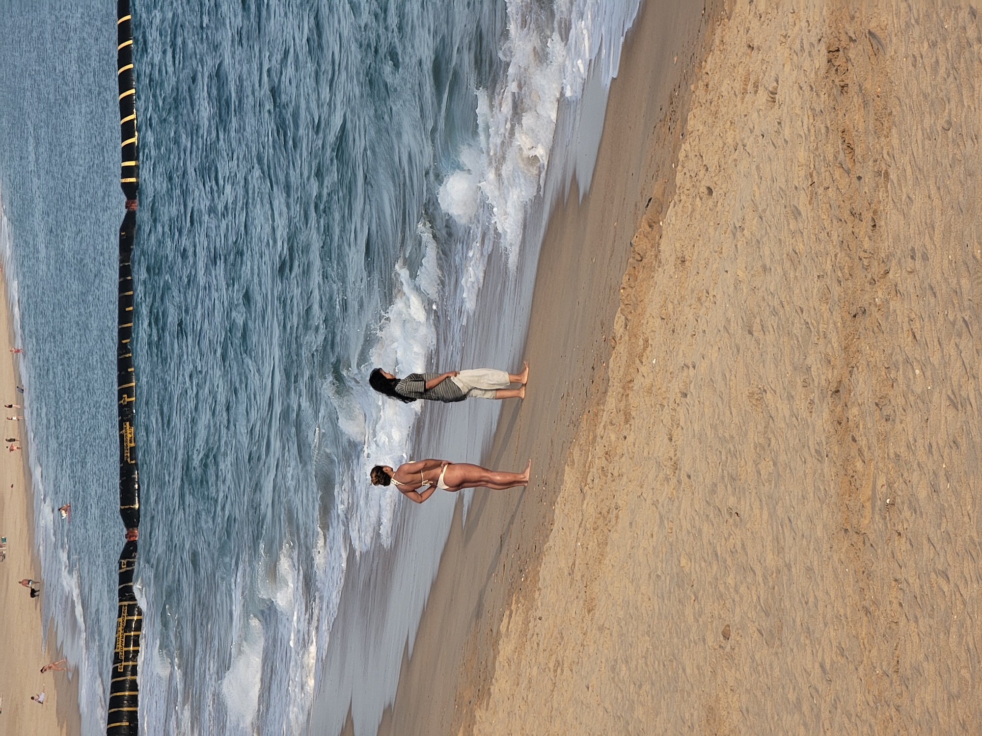 The image depicts a sandy beach in Quarteira, Portugal, with the Atlantic Ocean in the background. Two women are standing on the wet sand near the water's edge. The woman on the left is wearing a white bikini and is facing away from the camera with her hands on her hips. The woman on the right is wearing a striped shirt and light-colored pants and is facing towards the ocean. The water is blue and choppy, with waves breaking on the shore. A yellow and black barrier is visible in the ocean in the middle ground. The sand in the foreground is dry and textured with visible footprints. The sky is not visible.
