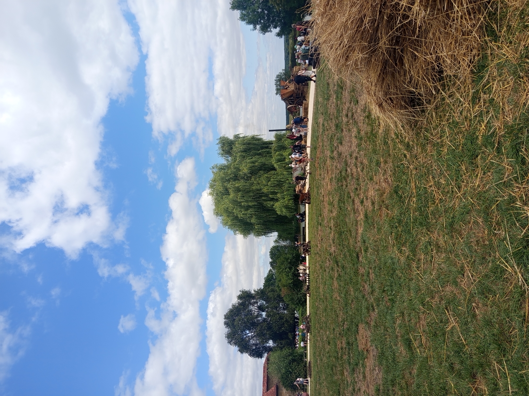 The image depicts an outdoor event or festival at the Parc de yesterday in Azannes-et-Soumazannes, France. In the foreground, a large pile of hay sits on a grassy field. In the middle ground, a group of people are gathered, observing various historical agricultural machines and equipment displayed along a dirt path. Several large trees, including a prominent weeping willow, and a building with a red-tiled roof are visible in the background. The sky is blue with scattered white clouds.
