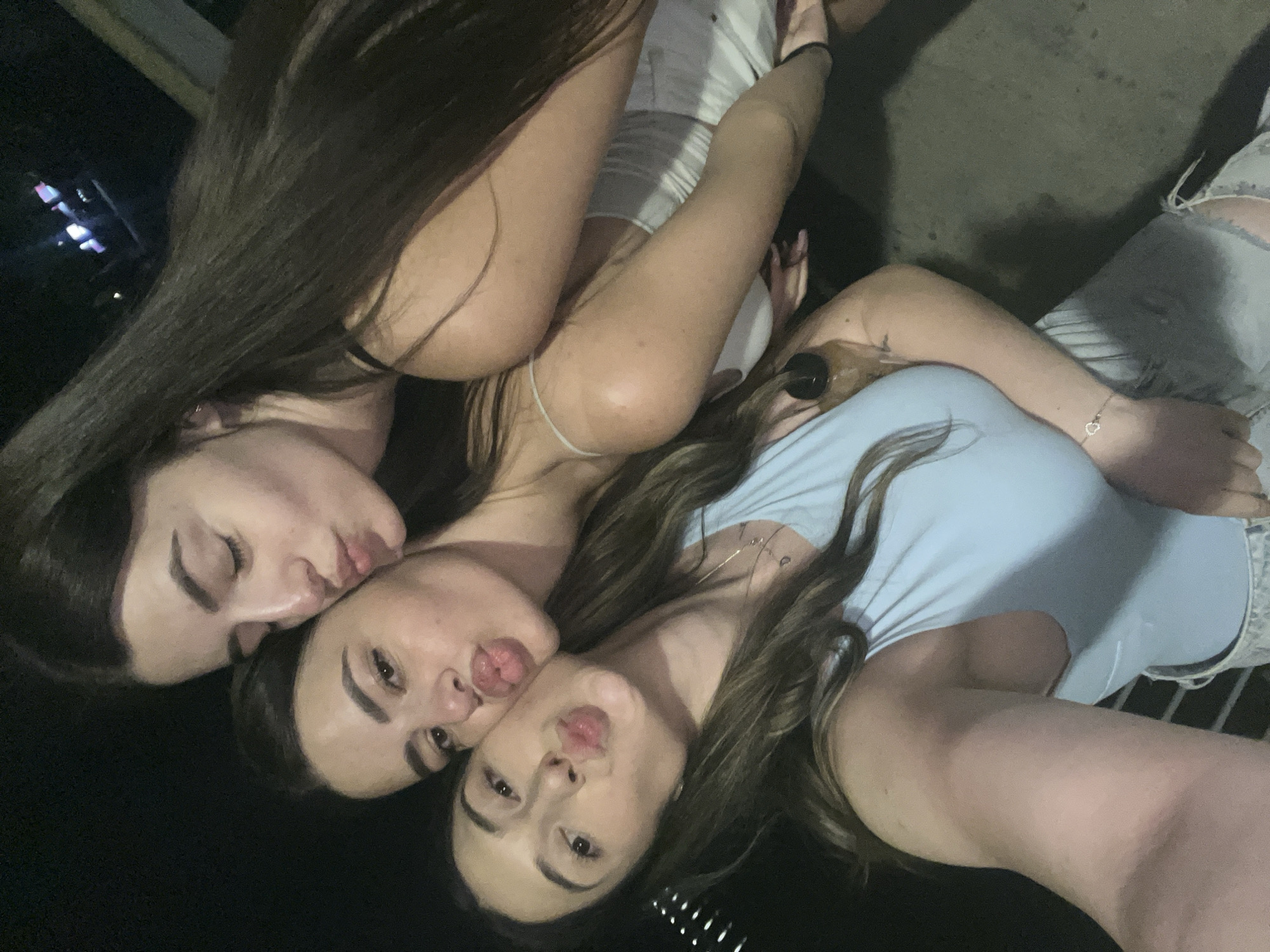 In a nighttime, outdoor setting, three young women are taking a selfie. The woman on the left, with long brown hair, is wearing a light blue top and distressed denim shorts. She has a delicate bracelet with a heart charm on her left wrist. The woman in the middle, also with long brown hair, is wearing a white tank top. The woman on the right, with dark brown hair, is leaning in, with her eyes closed. All three women are puckering their lips as if for a kiss. The background is dark, with some blurry lights visible in the distance, suggesting an urban environment. The scene is likely in Plovdiv, Bulgaria, given the prompt.