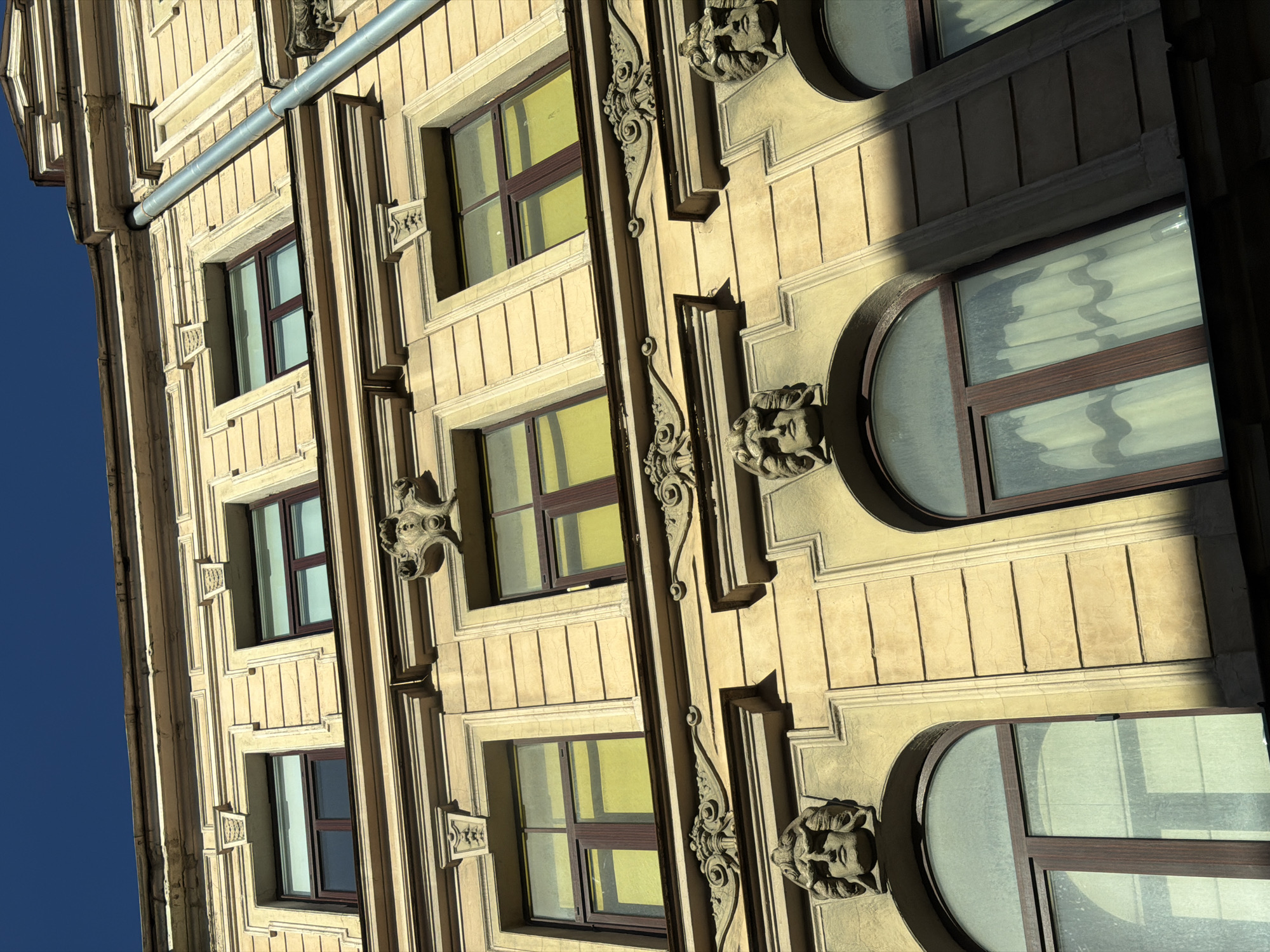 The image shows the facade of a building in Riga, Latvia, taken from a low angle. The building is several stories tall and has a light yellow, textured exterior. It is decorated with architectural details such as rectangular and arched windows, decorative moldings, and sculptural elements featuring faces between windows. A dark blue sky is visible in the upper-left corner. The lighting indicates it is daytime, with strong shadows cast on the building's surface.