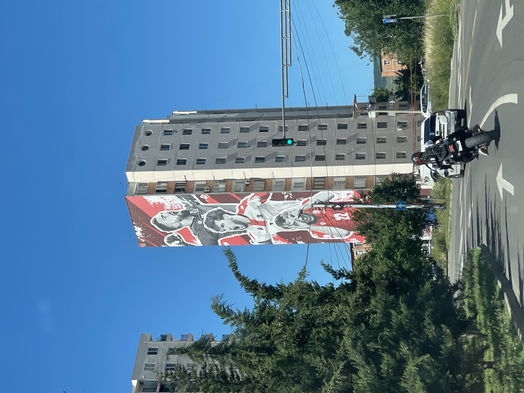 The image shows a tall residential building in Tbilisi, Georgia, adorned with a large mural of three football players in action. The mural features players in white and red jerseys with the number 22 visible on the lower player's jersey. One player is in the foreground, mouth open in a shout, while two others are depicted above him, also in dynamic poses. The building itself is a typical Soviet-era block structure, light grey with multiple windows and balconies.
In the foreground, a motorcycle with a rider wearing a black jacket and helmet is captured from behind, traveling on a multi-lane road. The road has white directional arrows painted on it, indicating traffic flow. To the left of the road, a large coniferous tree with lush green foliage is prominent. Across the street, more trees and grassy areas are visible, along with other buildings and a traffic light showing green. The sky is a clear, bright blue. The scene depicts a sunny day in an urban environment.