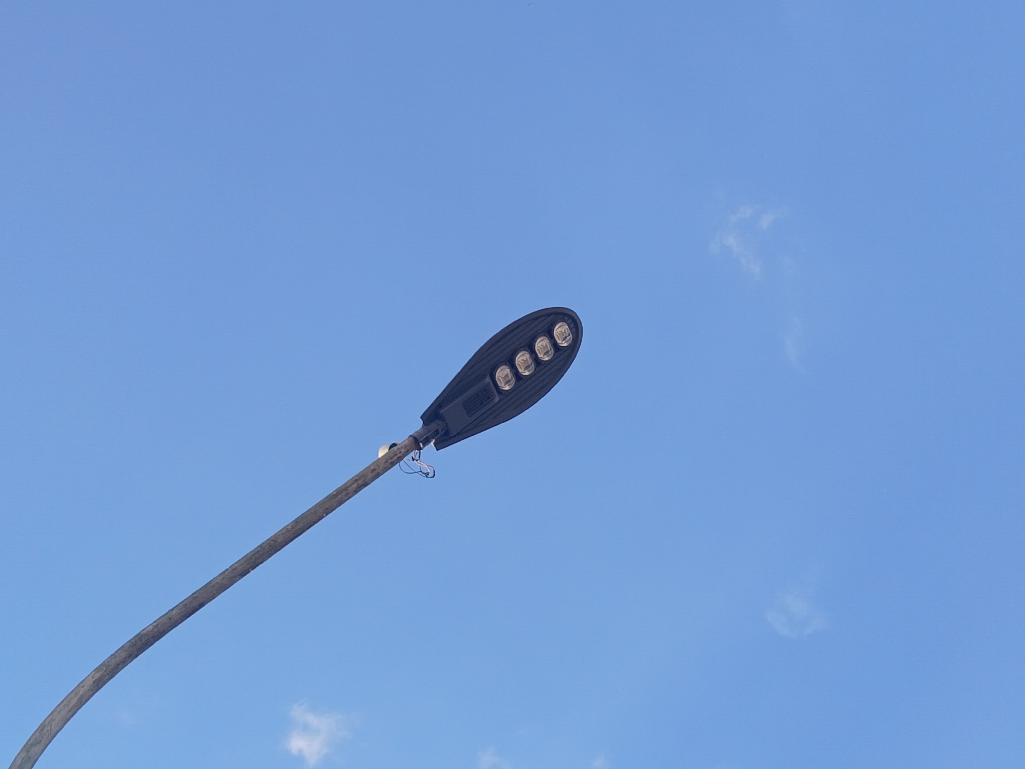 The image shows a streetlight in Guayana City, Venezuela against a clear blue sky with a few wisps of clouds. The streetlight is a dark color, possibly black or dark gray, and features five LED lights. The pole supporting the light is curved. There are some wires near where the light fixture is connected to the pole.