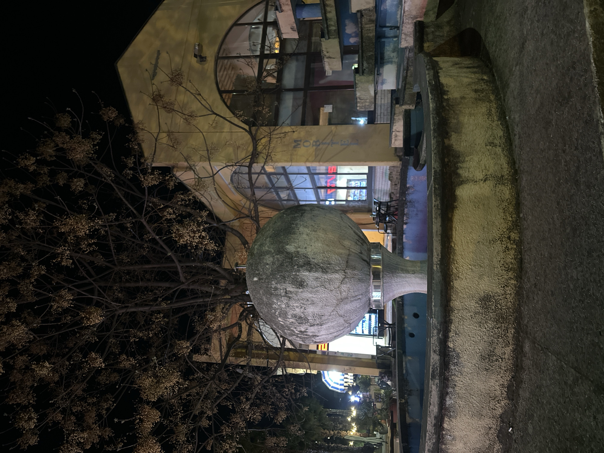 The scene depicts an urban public space at night in Budva, Montenegro. A large, spherical, light gray concrete or stone fountain dominates the midground, positioned on a conical pedestal within a circular basin. The basin is empty of water, featuring a faded blue interior and a discolored concrete exterior wall. The immediate foreground consists of dark, textured pavement. Behind the fountain, a two-story building with a light yellow-ochre facade is visible. This building features prominent arched windows on its upper level and rectangular windows on the lower, reflecting interior lighting. The word "MOBITEL" is vertically painted on the building's facade to the right of the central archway. Adjacent to the building on the right, a multi-tiered concrete structure, possibly a decorative cascade or steps, exhibits patches of faded blue and white paint. To the left of the fountain, a large deciduous tree, mostly bare, displays clusters of small, dried seed pods or foliage against the dark night sky. In the background to the left, other illuminated commercial structures, including one with a distinct blue and white striped awning, are discernible. The area is lit by artificial light sources. No individuals are present in the image. The fountain and surrounding concrete elements show signs of weathering and discoloration.