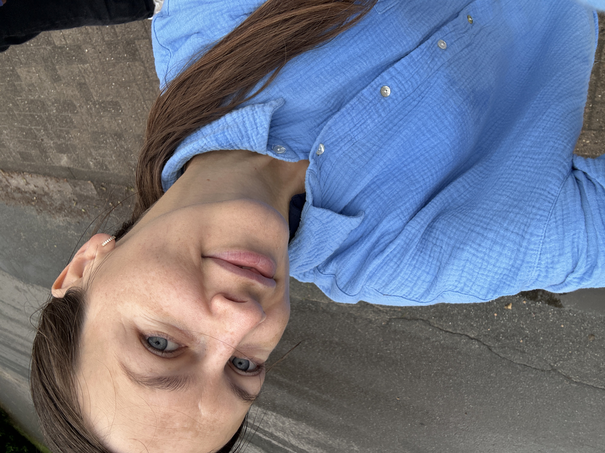 A selfie-style portrait captured outdoors in Deinste, Germany, features a woman with light blue eyes and long, medium-brown hair pulled back, visible over her left shoulder. She exhibits a subtle, closed-lip smile and appears to wear minimal makeup, with visible freckles or sunspots on her face. Her right earlobe displays a small, embellished hoop earring. She is wearing a light blue, textured, collared shirt with visible buttons down the front. The image is taken from a slightly elevated perspective, looking down. The background primarily consists of dark, textured ground, consistent with asphalt or concrete, showing some cracks and granular details. A darker, indistinct area, possibly foliage or shadow, is visible in the upper left quadrant.