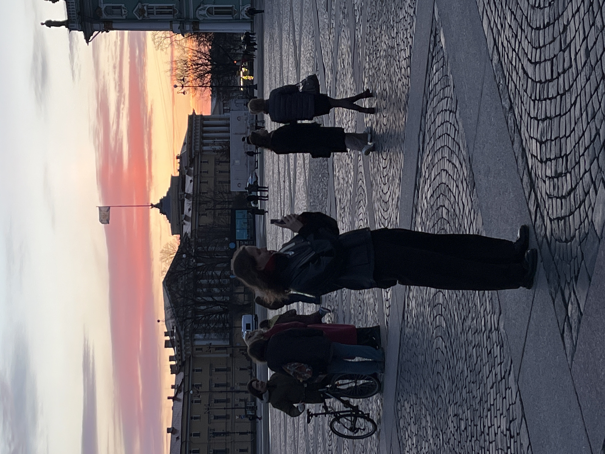 The image shows a group of people in Palace Square, Saint Petersburg, Russia, at sunset. A woman in a black jacket is holding up her phone, presumably taking a picture of the colorful sky. Several other people are walking in the background. There is a bicycle on the right, with the rider and the bicycle are in the foreground. The architecture of the buildings lining the square is visible in the background. The pavement is made of cobblestones.