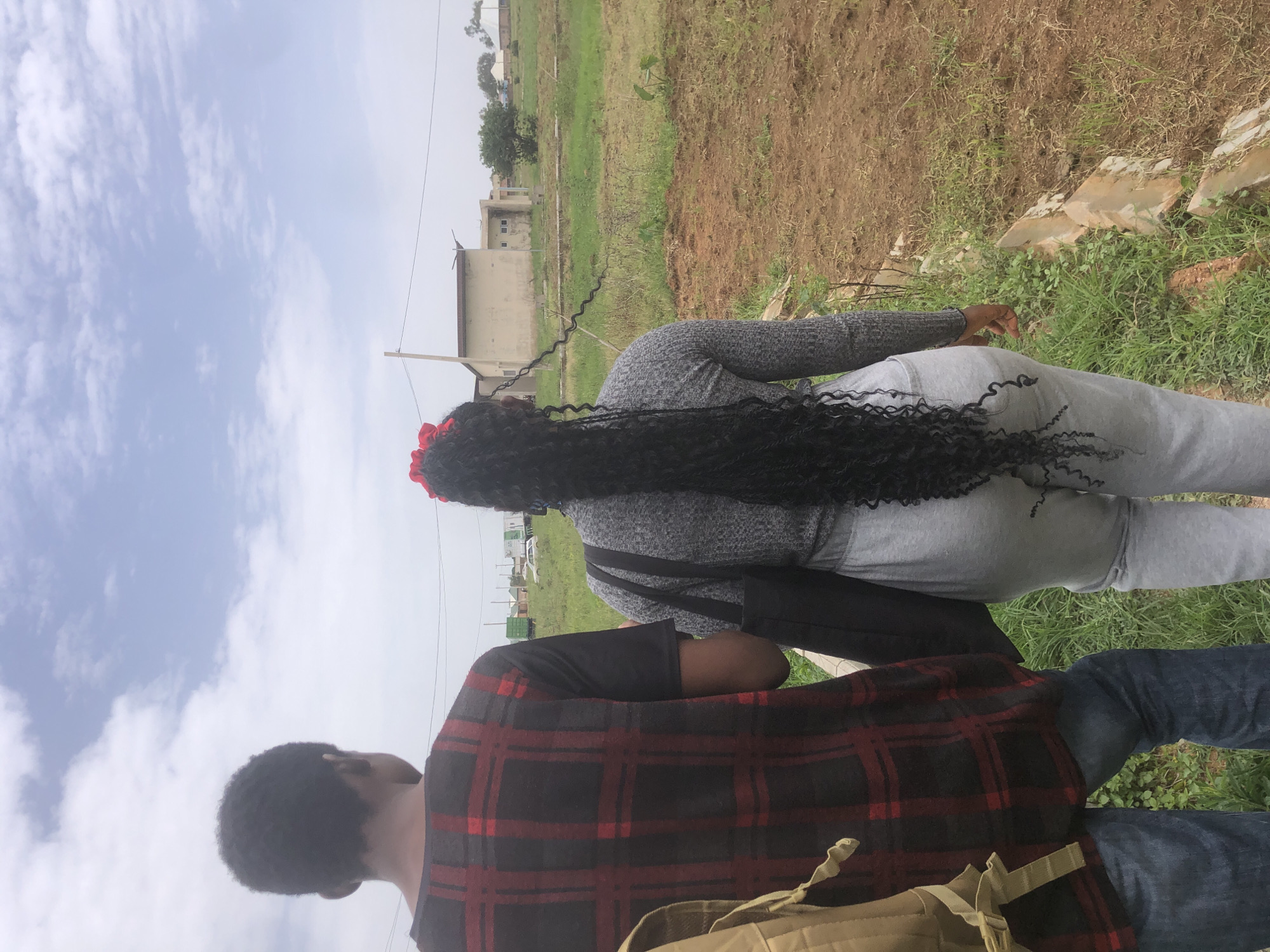 The media file shows two people from behind in an outdoor, rural setting in Gwafan, Nigeria. The person in the foreground is wearing a red and black plaid shirt and blue jeans, with a tan backpack visible at the bottom of the frame. To their left and slightly behind is another person with long, dark, curly hair adorned with a red accessory. This individual is wearing a grey sweater and grey pants, and a black bag is slung over their shoulder.
The background consists of undeveloped land with patches of dry earth and green grass, suggesting a dry season or an area undergoing construction. Some buildings, possibly houses or unfinished structures, are visible in the distance, along with some trees and vegetation. The sky is cloudy, indicating a possibly overcast or partly sunny day. The overall impression is of a casual outing or a moment of rest outdoors. There is no visible text in the media file.