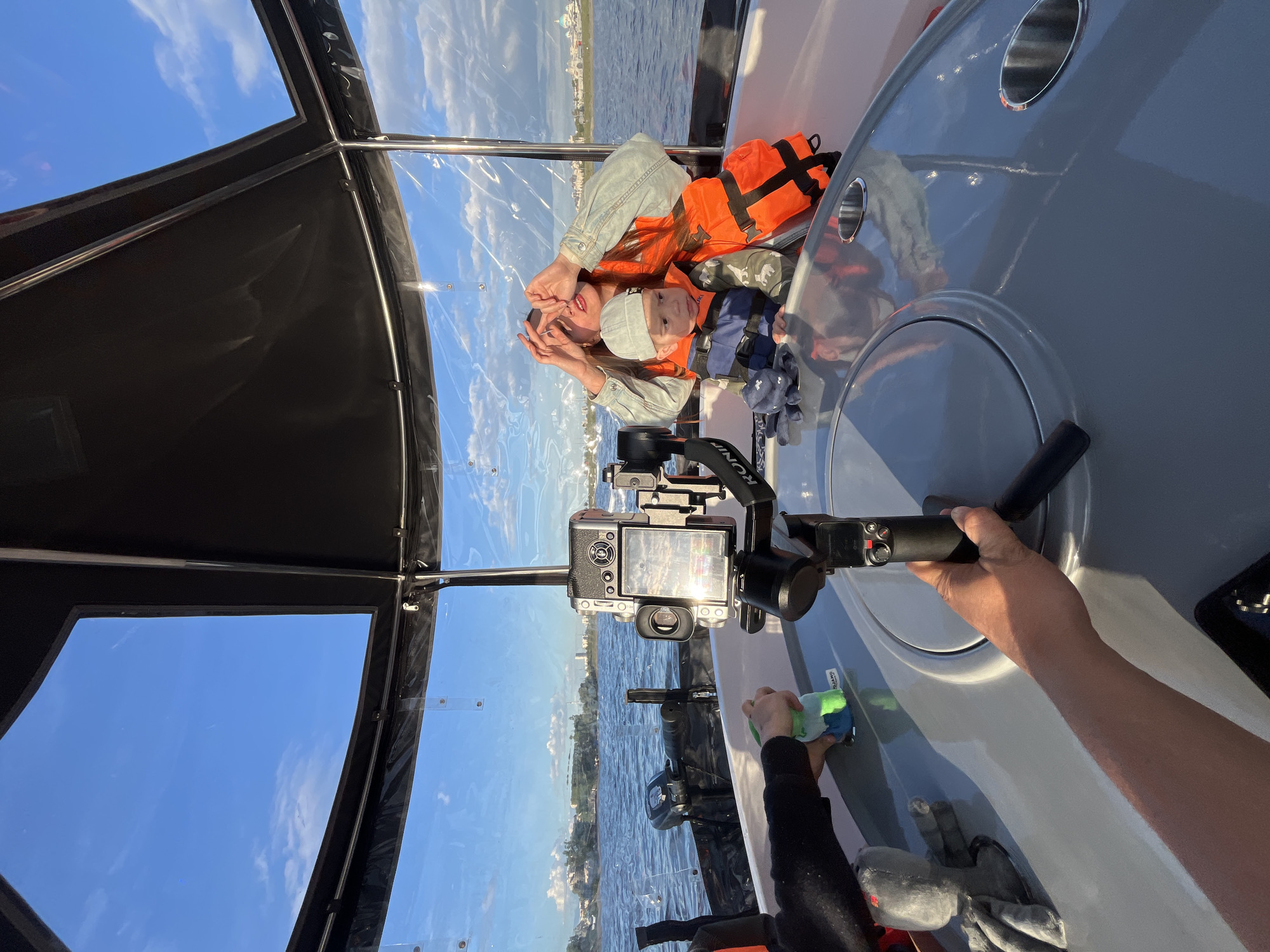 The image shows a woman and a child in a boat on the Kazanka River in Kazan, Russia. The woman is wearing a light-colored denim jacket and the child is wearing an orange life jacket and a gray hat. They are both looking at a camera that is mounted on a gimbal. The gimbal is attached to a boat's console, and an arm can be seen holding a cleaning rag, likely wiping the console. The boat has a clear plastic enclosure. The sky is blue with some clouds. The river is also visible through the enclosure. The cityscape of Kazan is partially reflected in the clear plastic.