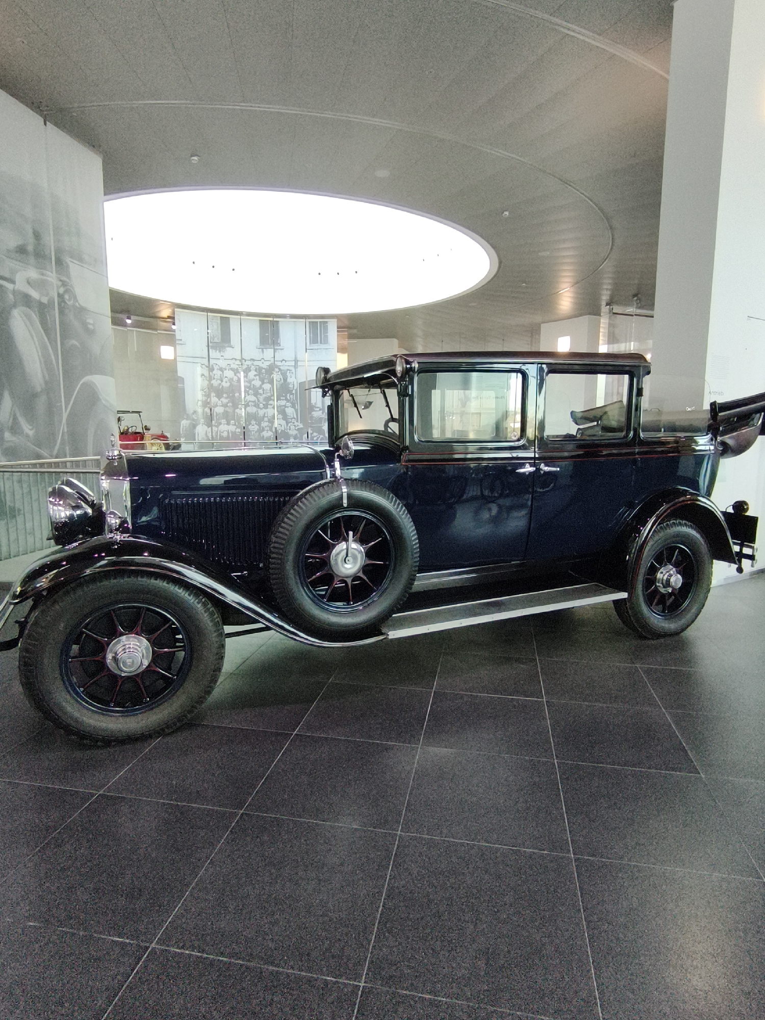 This is a full-length, eye-level, side-profile shot of a dark blue vintage automobile parked indoors on a dark tiled floor. The car appears to be a sedan with a spare tire mounted on the side, a tall grille, and chrome accents on the headlights and bumper. The vehicle is positioned in what appears to be a museum or exhibition space, indicated by the large, abstract ceiling structure with a circular skylight, a metal railing along the left side, and large photographic murals on the walls in the background. One mural depicts a black and white image of vintage cars, and another shows a group of people. A small red car can be seen in the distance behind the main subject. The lighting is bright and diffused, coming from the skylight and overhead fixtures. The overall impression is of a well-maintained exhibit in a modern architectural setting. This scene is likely located at the Audi Forum Ingolstadt in Ingolstadt, Germany, which houses a museum dedicated to the history of Audi.
