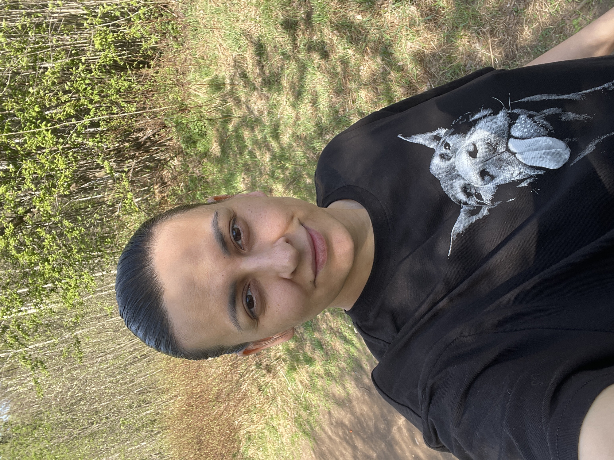 A person is shown in a close-up selfie outdoors. The person has short, dark hair slicked back and is wearing a black t-shirt with a white graphic of a dog. The background shows trees and grass. The location is not identifiable.