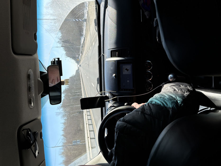 The image depicts the interior of a moving vehicle from a rear passenger's perspective, looking forward through the windshield onto a road in Spas-Zaulok, Russia. A driver is visible on the left, wearing a dark and teal puffer jacket, with their left hand gripping a black steering wheel. A manual transmission gear stick with a silver knob is visible on the center console. A black smartphone is mounted vertically on the dashboard and connected by a white cable. The vehicle's infotainment screen displays a music interface with the text "МУЗЫКА" and an album art icon. Above the windshield, dark folded sunglasses are clipped to the overhead console, and a rearview mirror reflects the lower half of a person's face. A black dashcam is mounted to the right of the rearview mirror, and a small, rectangular air freshener hangs from the mirror stem. The windshield is noticeably dirty or smudged, particularly on the right side.
Outside the vehicle, a multi-lane asphalt road with white markings extends into the distance under a clear blue sky. White metal guardrails line both sides of the road. The surrounding environment features bare trees, utility poles, and patches of snow on the ground along the roadside, suggesting a late winter or early spring setting. Distant vehicles are visible on the road ahead.