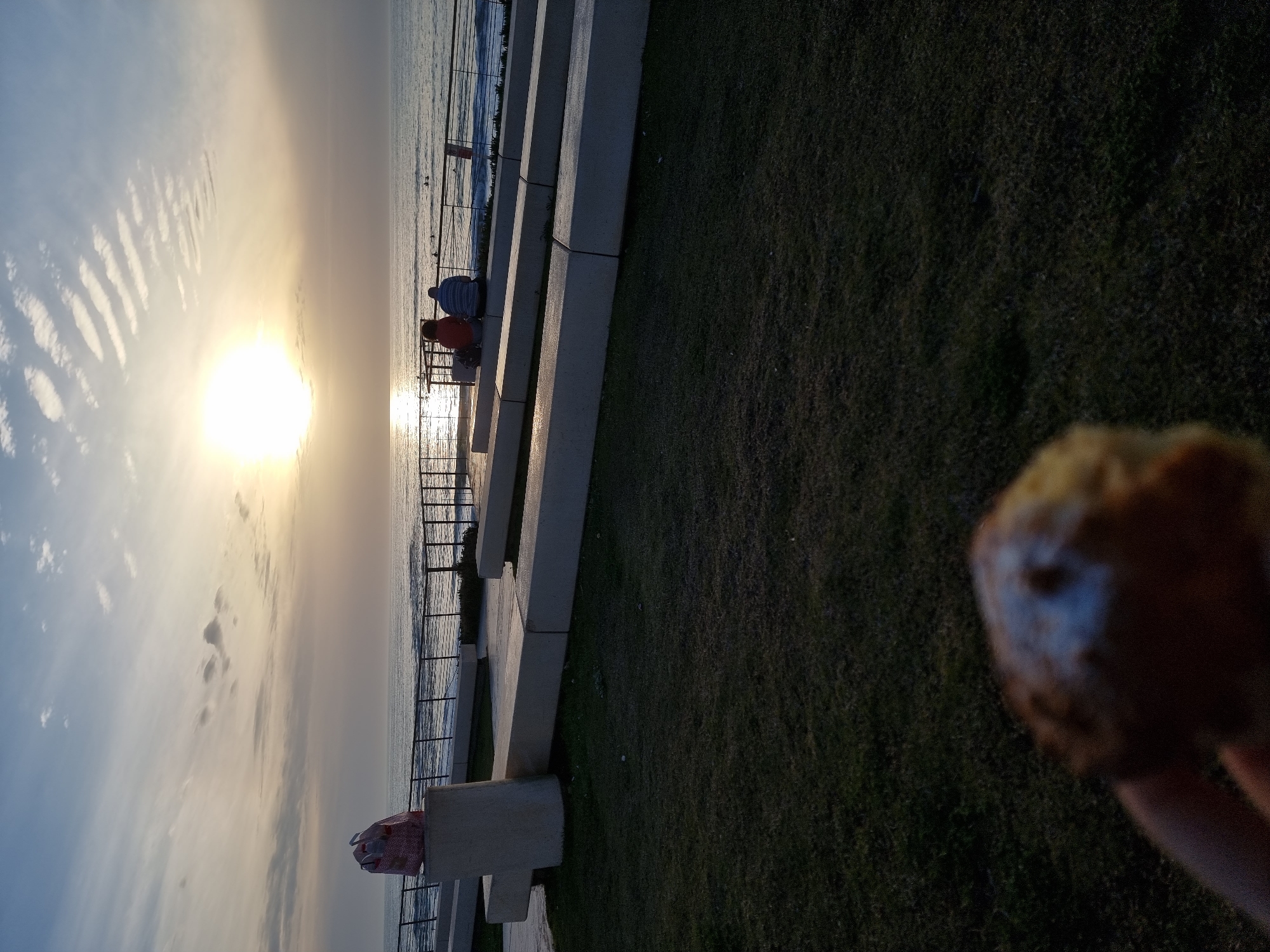 The image depicts a coastal scene along the Tel-Aviv promenade in Israel, observed at either sunrise or sunset. In the immediate foreground, a blurred hand holds a light-brown, circular pastry topped with white powdered sugar. The mid-ground features a dark green grass lawn bordered by light gray concrete tiered seating structures. On the left, a tall cylindrical concrete pillar is topped with a red and white patterned reusable bag. A dark metal railing separates the public seating area from the Mediterranean Sea. Two individuals are seated on the uppermost concrete bench, facing the sea. One wears a reddish-orange top and has a dark backpack or bag beside them; the other wears a blue and white striped shirt. In the background, the sun, a bright circular orb, is low on the horizon, casting a strong reflection across the moderately wavy sea. Multiple small dark figures, consistent with surfers or swimmers, are visible in the water. The sky is partially cloudy, displaying distinct parallel wave-like cloud formations in the upper left.