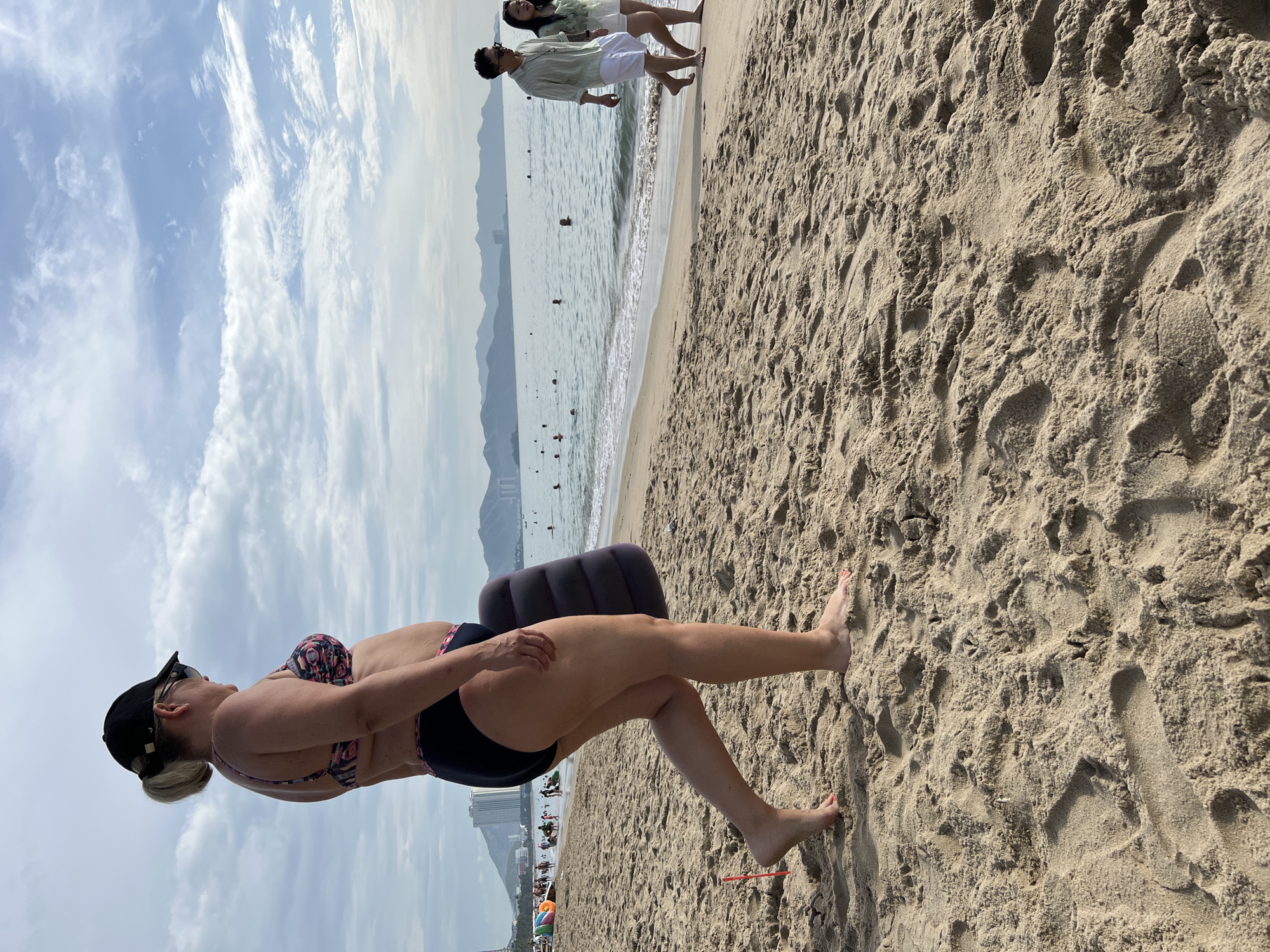 An adult Caucasian female wearing a black baseball cap, sunglasses, and a floral bikini walks on a sandy beach in Nha Trang, Vietnam. She is carrying an inflatable pillow. The ocean is to her right, with mountains in the background. A couple walks on the beach in the distance. The sky is cloudy and blue. The sand is light brown and textured, with footprints visible. In the background, buildings and palm trees are visible along the coastline.