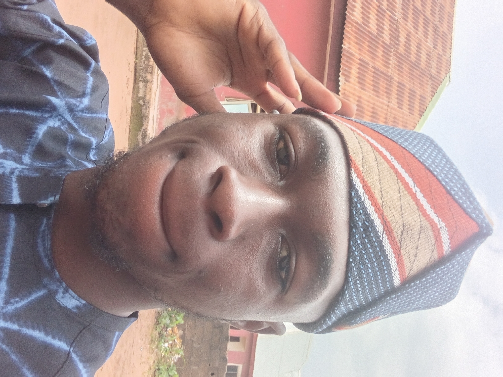 A close-up selfie shows a young Black man wearing a traditional Nigerian cap. He is outdoors, with a red-painted building and a corrugated metal roof visible in the background. The man is looking at the camera and has his left hand raised to his forehead, as if saluting or shading his eyes. He is wearing a dark blue and white patterned shirt. The sky is overcast. The overall setting appears to be a residential area in the city of Jalingo, Nigeria.