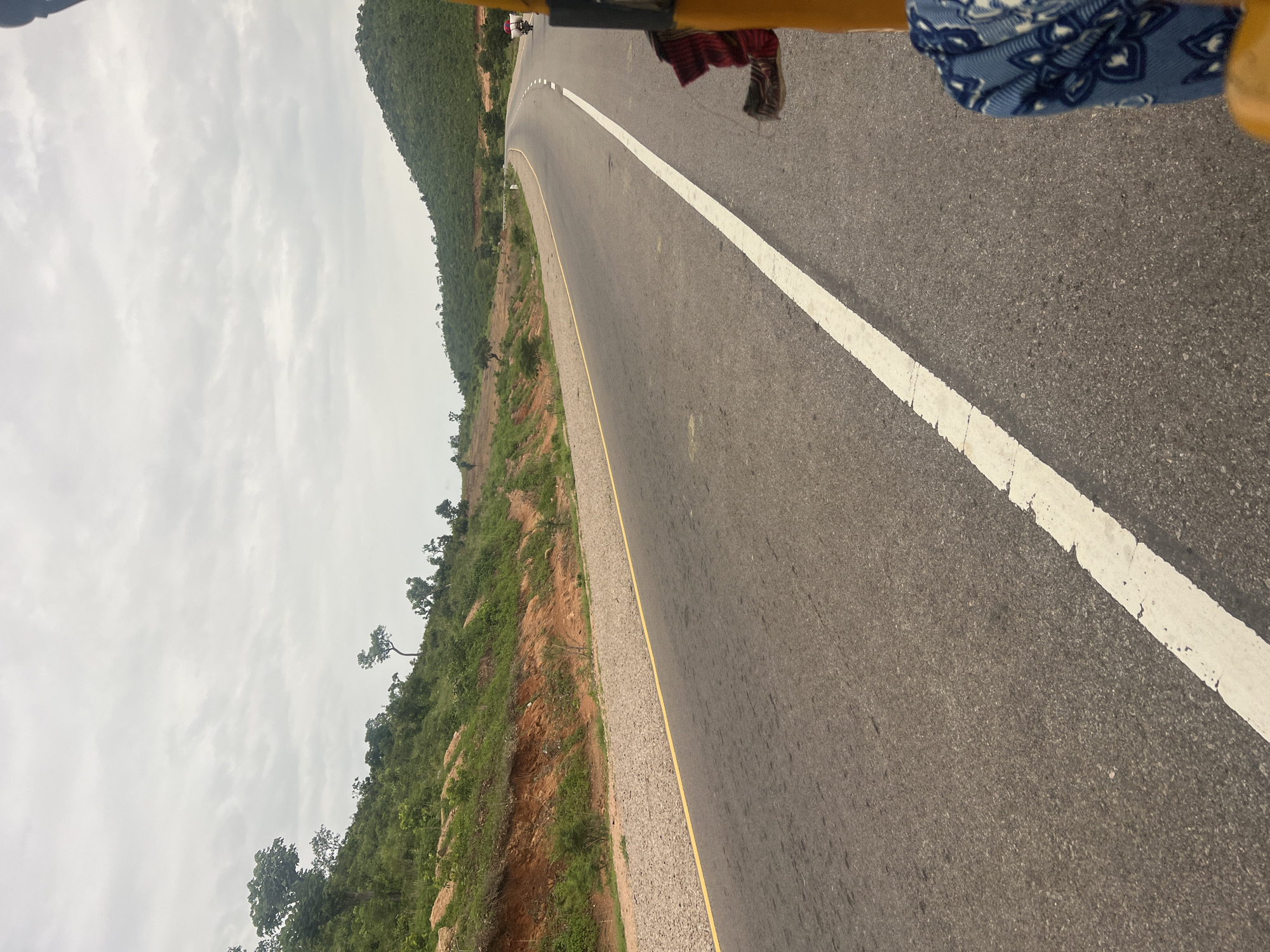 The image captures a view from a vehicle, likely a tricycle given the yellow framing visible at the top. The road ahead curves gently to the left, bordered by a white line on the right and a yellow line on the left. On the left side of the road, a lush green, tree-covered hillside rises steeply. The hillside is interspersed with patches of exposed red earth and rocks, suggesting a natural, somewhat rugged terrain. A few scattered trees stand out against the dense greenery. The sky is overcast with a layer of pale gray clouds, indicating a likely dull or rainy day. The overall mood is one of travel through a natural landscape. The scene appears to be a roadside view, with no specific landmarks that would pinpoint an exact location within Minda, Nigeria, beyond it being a road traversing a hilly, vegetated area. The time of day is indeterminate due to the overcast sky, but it appears to be daytime. There is no visible text in the image.