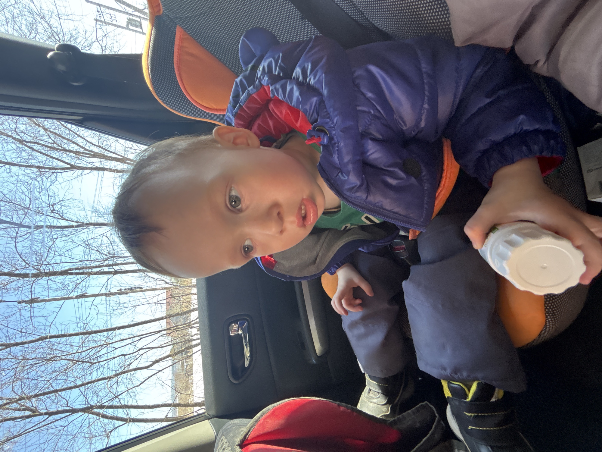 A toddler sits in a car seat in a vehicle. The child wears a puffy blue jacket and gray pants with black shoes featuring yellow trim. The child holds a white container. Trees without leaves are visible through the car window. The location is most likely not Ферма, Russia, as this cannot be determined from the image.
