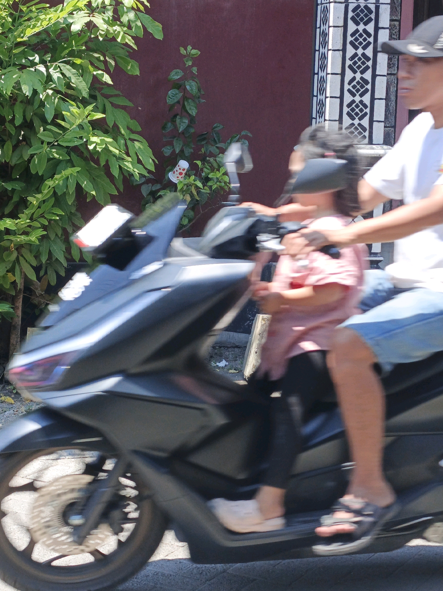 A motion-blurred image captures a man and a child on a motorcycle on a street in Ketegan, Indonesia. The man, wearing a white t-shirt and blue jeans, is driving. The child, in a pink shirt, sits in front of him. Green foliage is visible to the left of the motorcycle, and a building with a black and white tiled pillar is in the background. The motorcycle is dark gray with a silver front wheel.