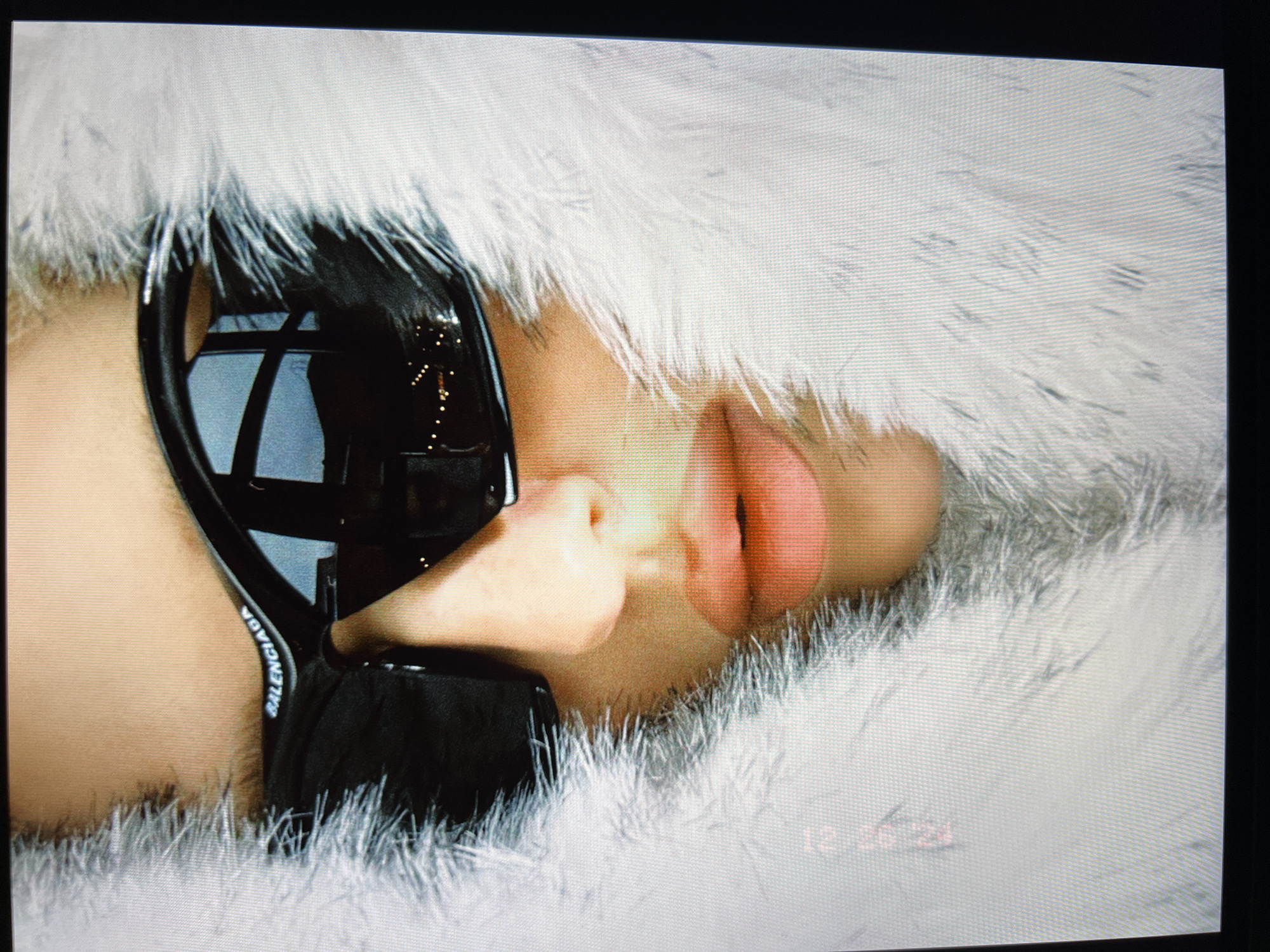 The image is a close-up, medium-shot photograph of a person wearing large, black, wraparound sunglasses with the "BALENCIAGA" logo visible on the arm. The person's face is framed by a thick, white, furry material, possibly a coat or scarf. The background is out of focus, but reflections in the sunglasses suggest an indoor setting with some lights. There is a timestamp in red text in the bottom left corner of the image that reads "18:24". The photograph was taken in Saint Petersburg, Russia.