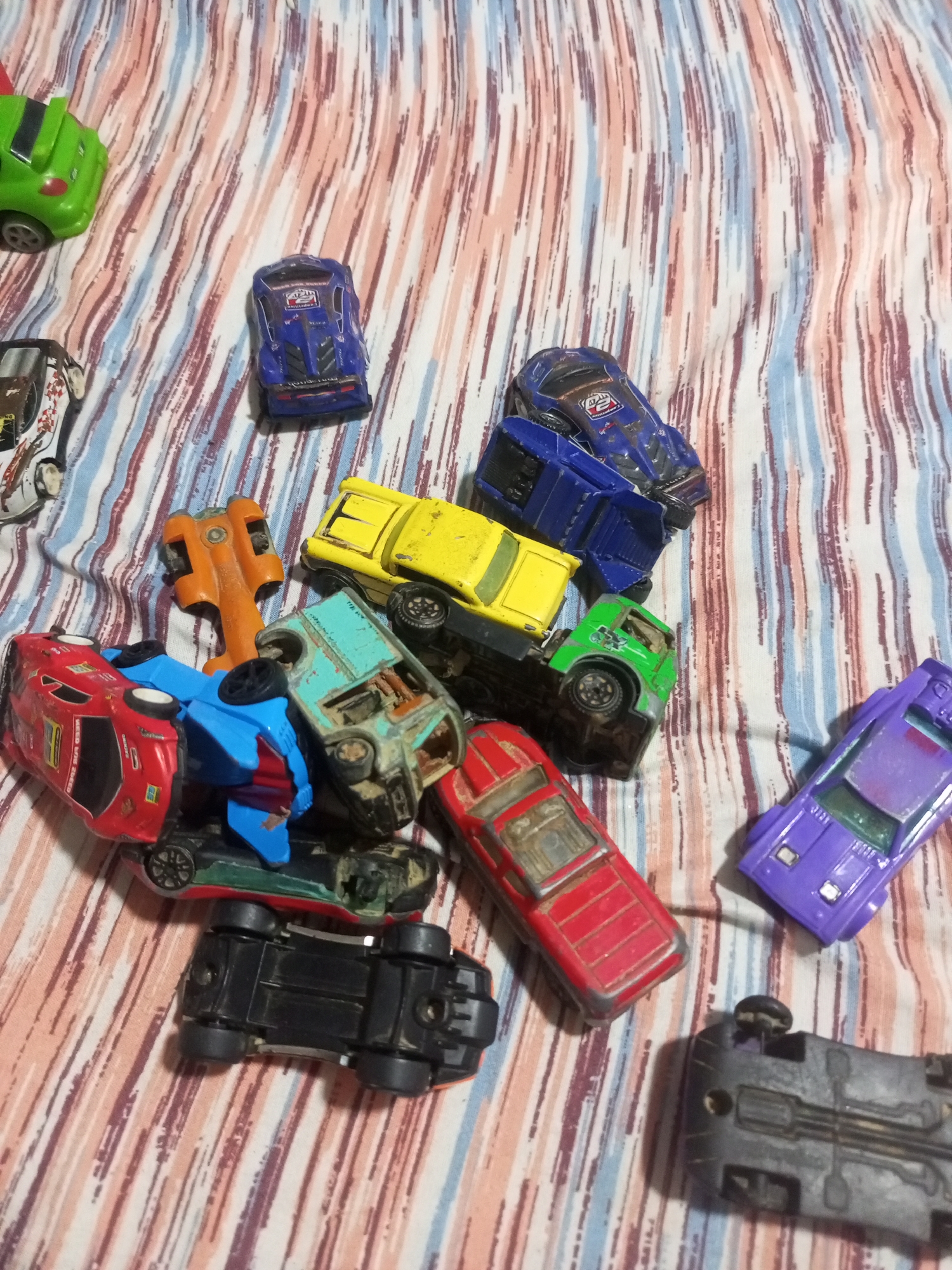 The image depicts a collection of toy cars scattered on a striped fabric surface. The fabric has vertical stripes in shades of pink, blue, and white. The toy cars are of various colors and types, including a green car, a blue race car, a yellow truck, an orange vehicle, a red car, a blue car, a green truck, and a purple car. Several cars are overturned or partially obscured by others. The scene appears to be indoors, with no visible elements suggesting a specific urban location like La Tagua, Venezuela. There are no people or discernible actions occurring in the image. The focus is solely on the arrangement of the toy cars.