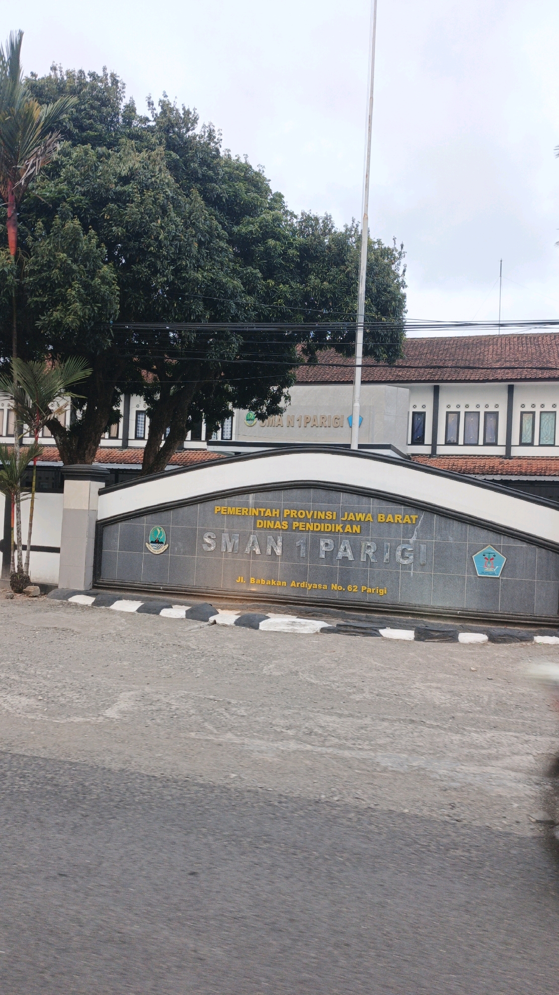 A medium shot captured from a slightly low angle shows the entrance to SMAN 1 Parigi, a school located in Desa Karangjaladri, Indonesia. The school's name is prominently displayed on a large, dark gray, curved sign with gold lettering. Above "SMAN 1 PARIGI," smaller yellow text reads "PEMERINTAH PROVINSI JAWA BARAT DINAS PENDIDIKAN." Below the school name, in smaller yellow text, is the address "Jl. Babakan Ardiysa No. 62 Parigi." Two school crests are visible, one on the left and one on the right side of the sign. The school building is visible behind the sign, a two-story structure with white walls and a brown tiled roof. Rows of windows with black frames are visible on the upper floor, and some decorative architectural elements are present. To the left of the entrance sign, part of a palm tree and a tall, slender palm-like tree are visible. The entrance to the school is framed by a stone pillar and a darker gray wall. The foreground shows a paved road with a black and white striped pedestrian crossing. The sky is overcast and a tall flagpole stands to the right of the school building. The overall scene depicts the entrance of an educational institution in a semi-urban or rural setting.