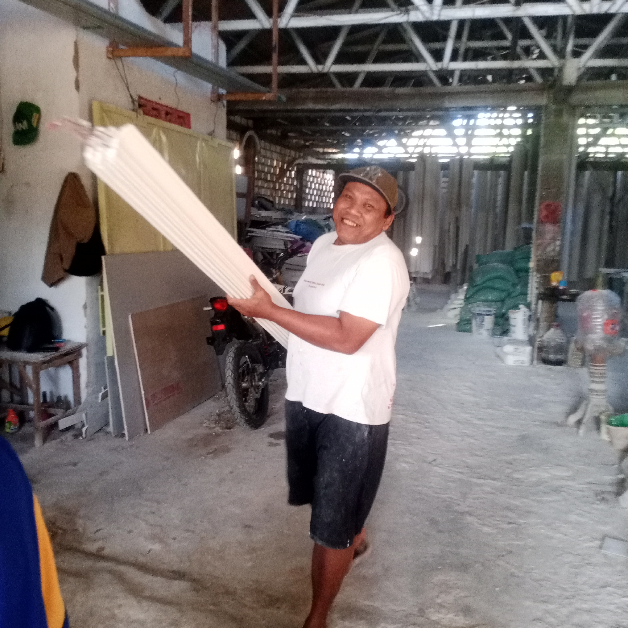In the city of Semanding, Indonesia, a person wearing a white t-shirt and dark shorts stands in what appears to be a storage or workshop area. They are smiling and holding a stack of long, white, flat objects, possibly building materials. The environment is dusty, with a concrete floor and a partially constructed ceiling. Various items are visible in the background, including a motorcycle, stacks of materials, and bags of what might be cement or fertilizer. The lighting suggests it is daytime.