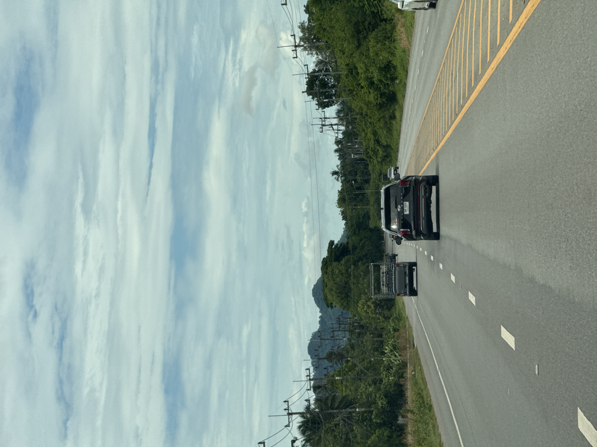 This is a roadside view on a cloudy day in Don Sak, Thailand. The image shows a paved road with a black SUV driving in the right lane, heading away from the camera. The SUV has a license plate that reads "SW 1035". To the left of the SUV, a silver pickup truck with a metallic cage bed is in the left lane, also heading away from the camera. Further ahead in the left lane, another vehicle is visible. The road is bordered by lush green vegetation and palm trees on both sides, with power lines and poles running along the left side. In the background, rolling green hills or mountains are visible under a sky filled with scattered clouds. The road markings include dashed white lines separating lanes and a solid yellow line with diagonal yellow markings on the right side of the right lane, possibly indicating a hazard or a breakdown area. The overall scene depicts a typical highway or main road in a tropical environment.