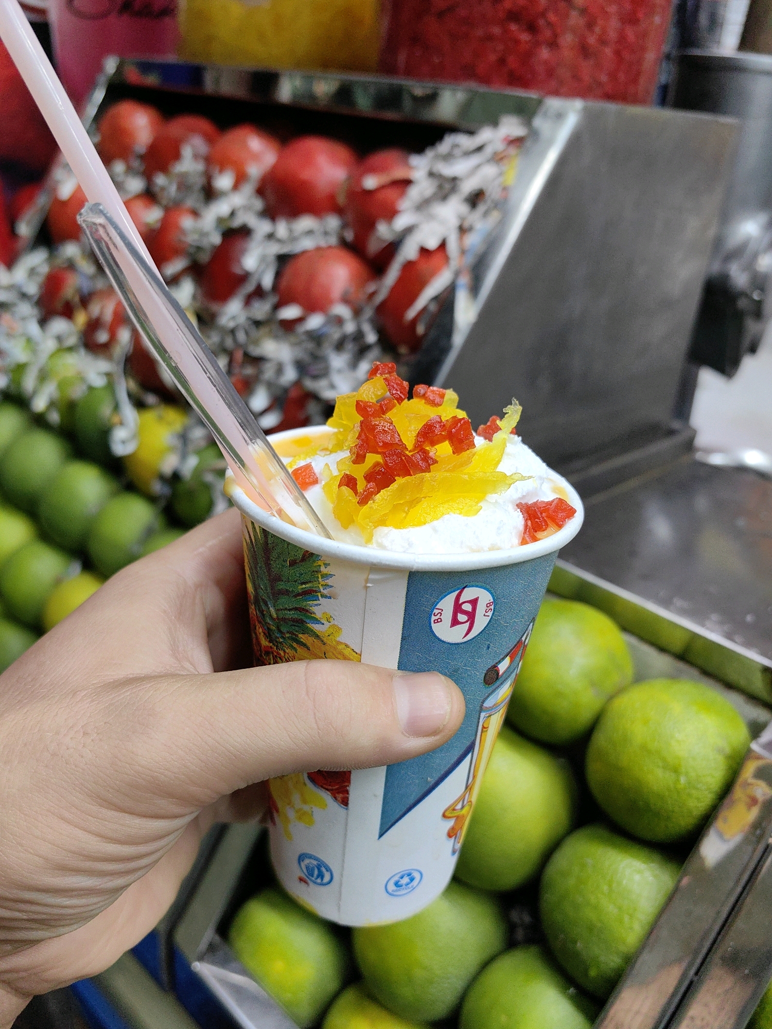 In Delhi, India, a person holds a paper cup filled with a creamy dessert, possibly a kulfi or ice cream, topped with chopped candied fruits. A plastic straw protrudes from the cup. The background reveals a vibrant display of fresh produce, including rows of green limes and what appear to be red tomatoes or apples, suggesting a market or street food stall. The overall scene is bright and colorful, indicative of a bustling Indian marketplace.