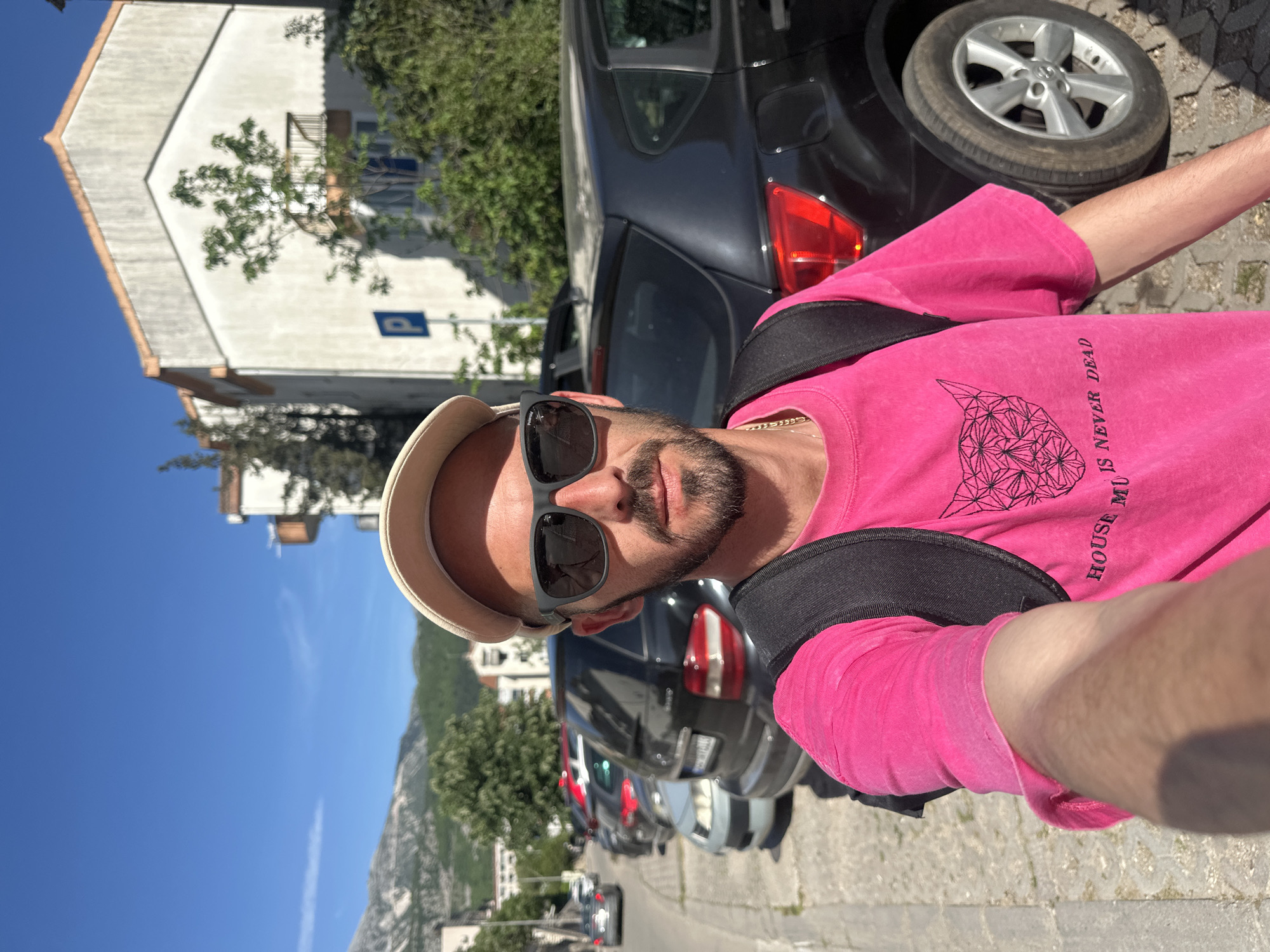 A bright, sunlit selfie captures a man from the chest up on a street in Budva, Montenegro. The man, with a beard and mustache, wears dark sunglasses, a light-colored cap, and a vibrant pink t-shirt. A black geometric design and partially visible text, "HOUSE MUSIC IS NEVER DEAD," are printed on his shirt. A gold chain is visible around his neck, and a black backpack strap crosses his shoulder.
The background reveals a bustling street scene. Behind the man, several cars are parked along what appears to be a stone-paved street. A prominent silver car wheel is visible close to the foreground. To the left, a multi-story white building with a distinctive peaked roof stands under a clear blue sky. A blue parking sign with a white 'P' is affixed to the building's facade. Green foliage from trees and bushes is scattered throughout the scene, adding touches of nature. In the distance, large mountains rise under the bright sky, with a faint contrail visible. The strong sunlight suggests it is daytime, casting clear illumination on the scene.