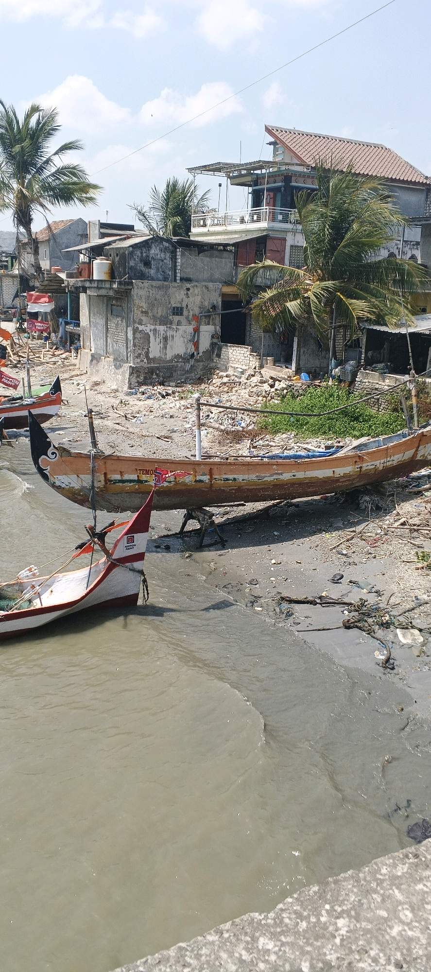 The image depicts a coastal scene in Tunggul, Indonesia, characterized by its weathered and somewhat dilapidated appearance. In the foreground, a murky body of water laps against a sandy and debris-strewn shore. Several fishing boats are present. One prominent boat, painted in orange and white with red accents, rests on the shore supported by wooden stands. The word "TEMOR" is visible on its side. Another smaller boat, predominantly white with red trim and a triangular flag bearing a red logo, is partially submerged in the water.
In the background, a collection of buildings forms the backdrop. These structures appear to be a mix of residential homes and possibly small businesses. They exhibit signs of age, with some walls showing exposed brickwork and debris scattered around. Palm trees are interspersed among the buildings, adding to the tropical ambiance. The sky above is a clear blue with scattered white clouds, suggesting a sunny day. The overall impression is one of a working fishing village, possibly experiencing economic hardship or recovering from a natural event, given the presence of scattered debris and the weathered state of the infrastructure.